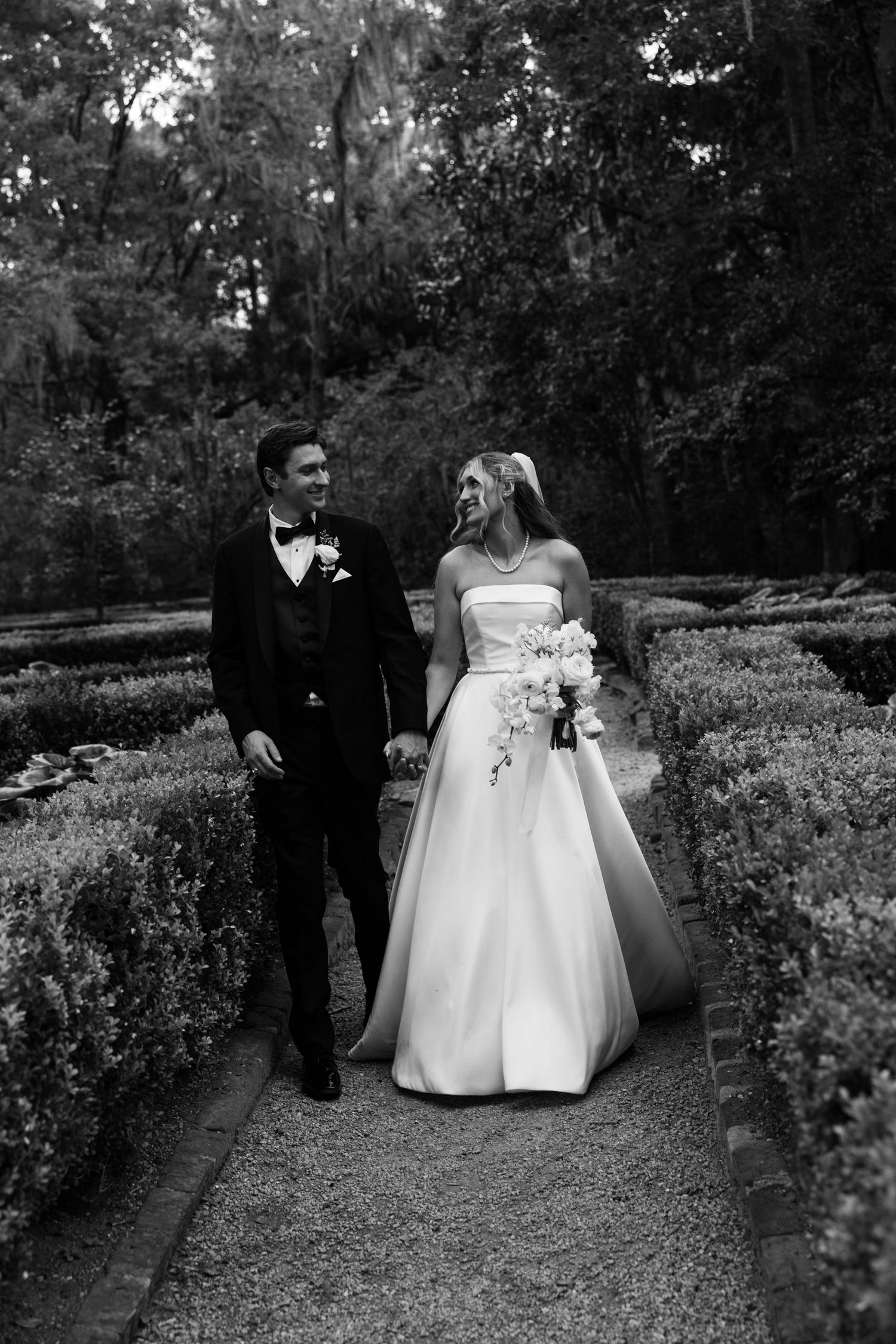 Black and white photo of a bride and groom walking hand in hand through a lush garden pathway on their wedding day.