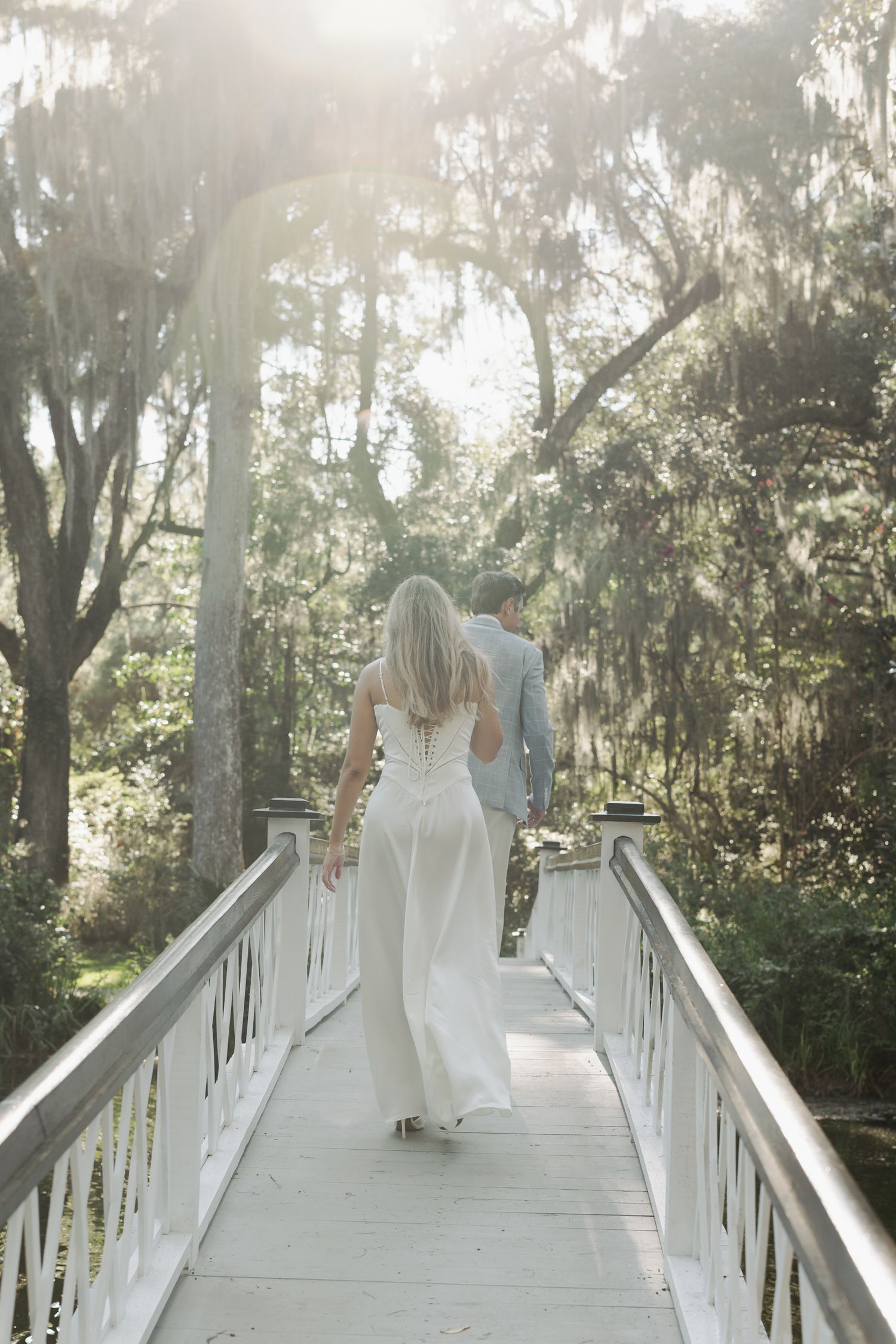 Bride and groom walking hand in hand across a white bridge surrounded by moss-draped trees in soft sunlight.