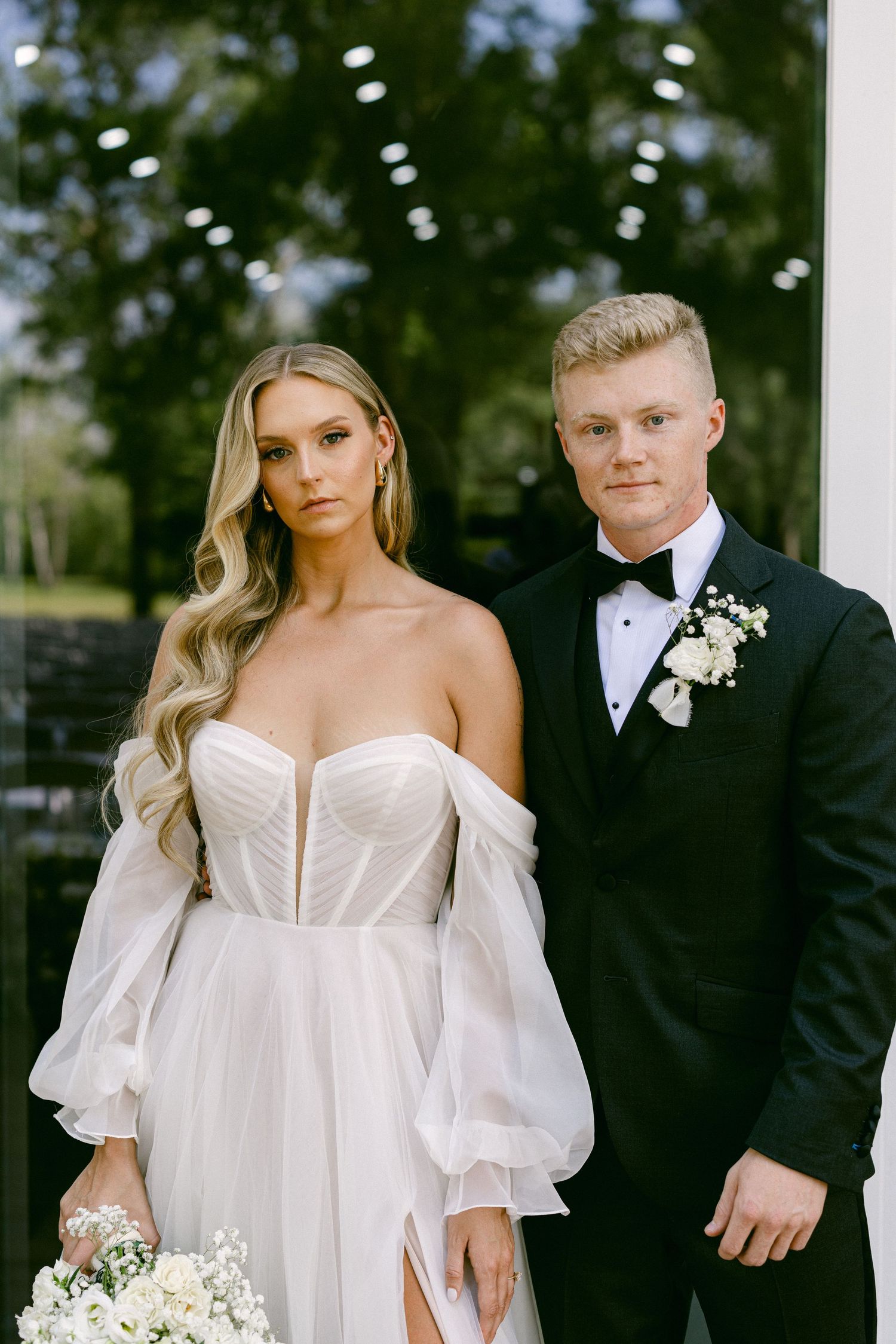 Elegant bride in off-shoulder white gown and groom in black tuxedo posing together at wedding.