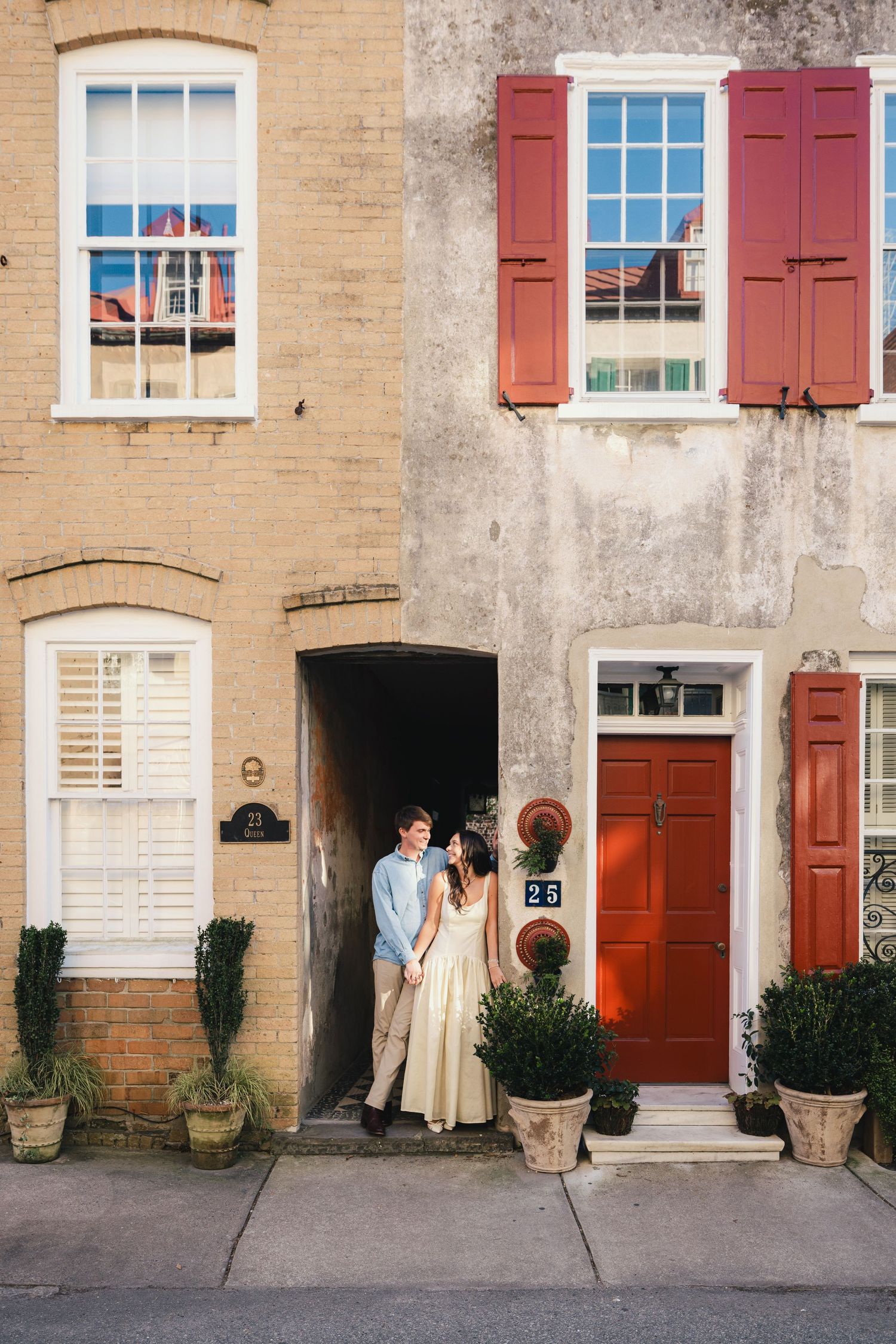 Couple standing in doorway of charming historic Charleston row houses with red shutters and potted topiaries.