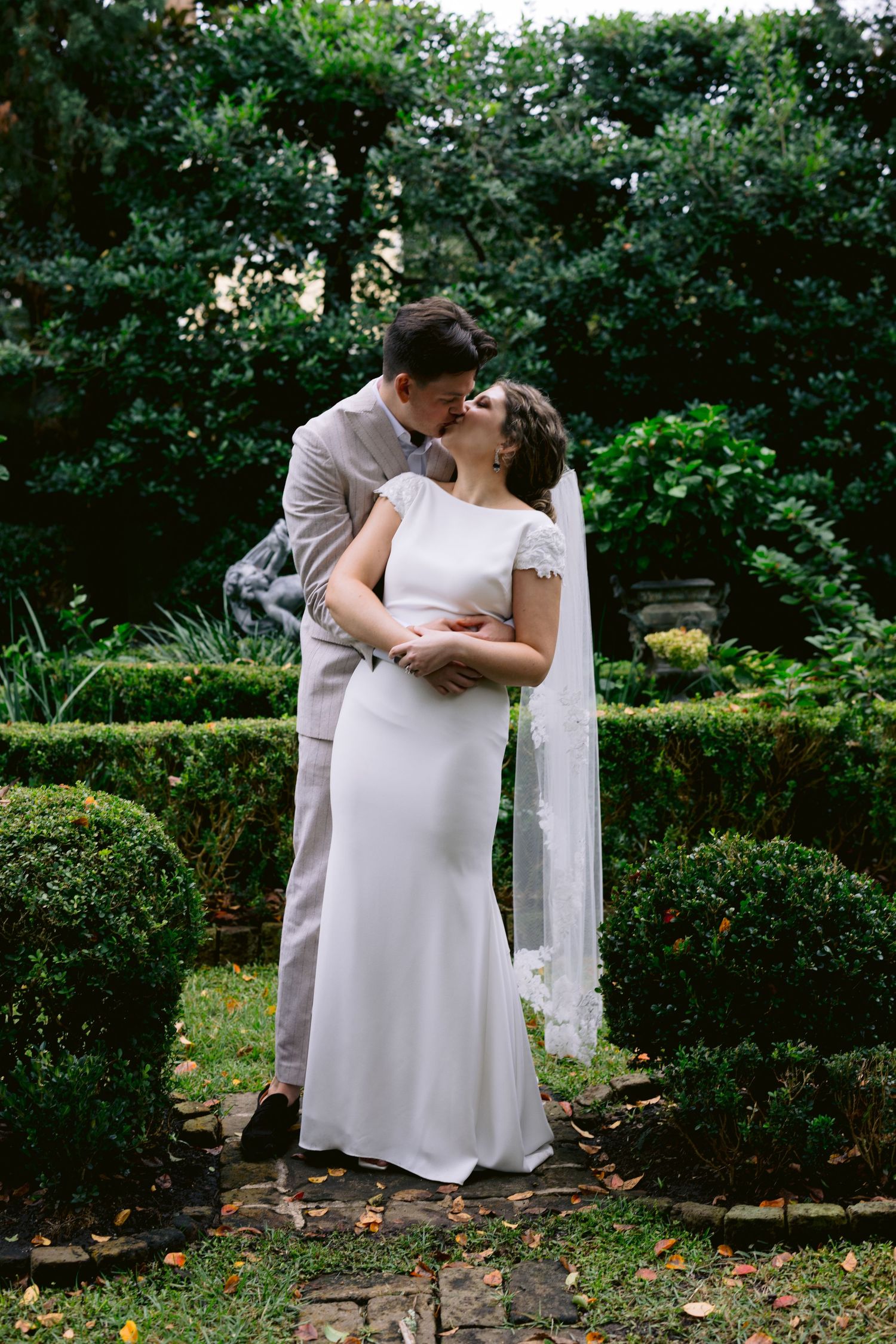 Bride and groom sharing a kiss in formal garden with manicured hedges and lush green backdrop.