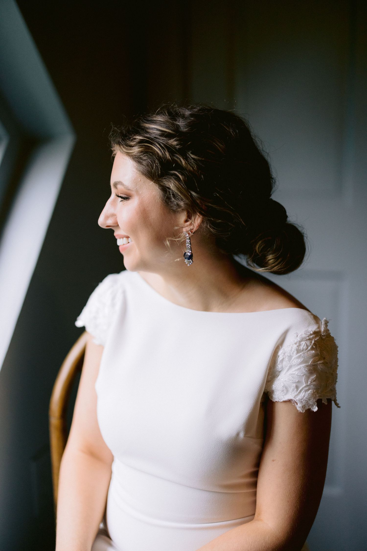 Radiant bride in white cap sleeve gown with lace detail and drop earrings smiling by window.