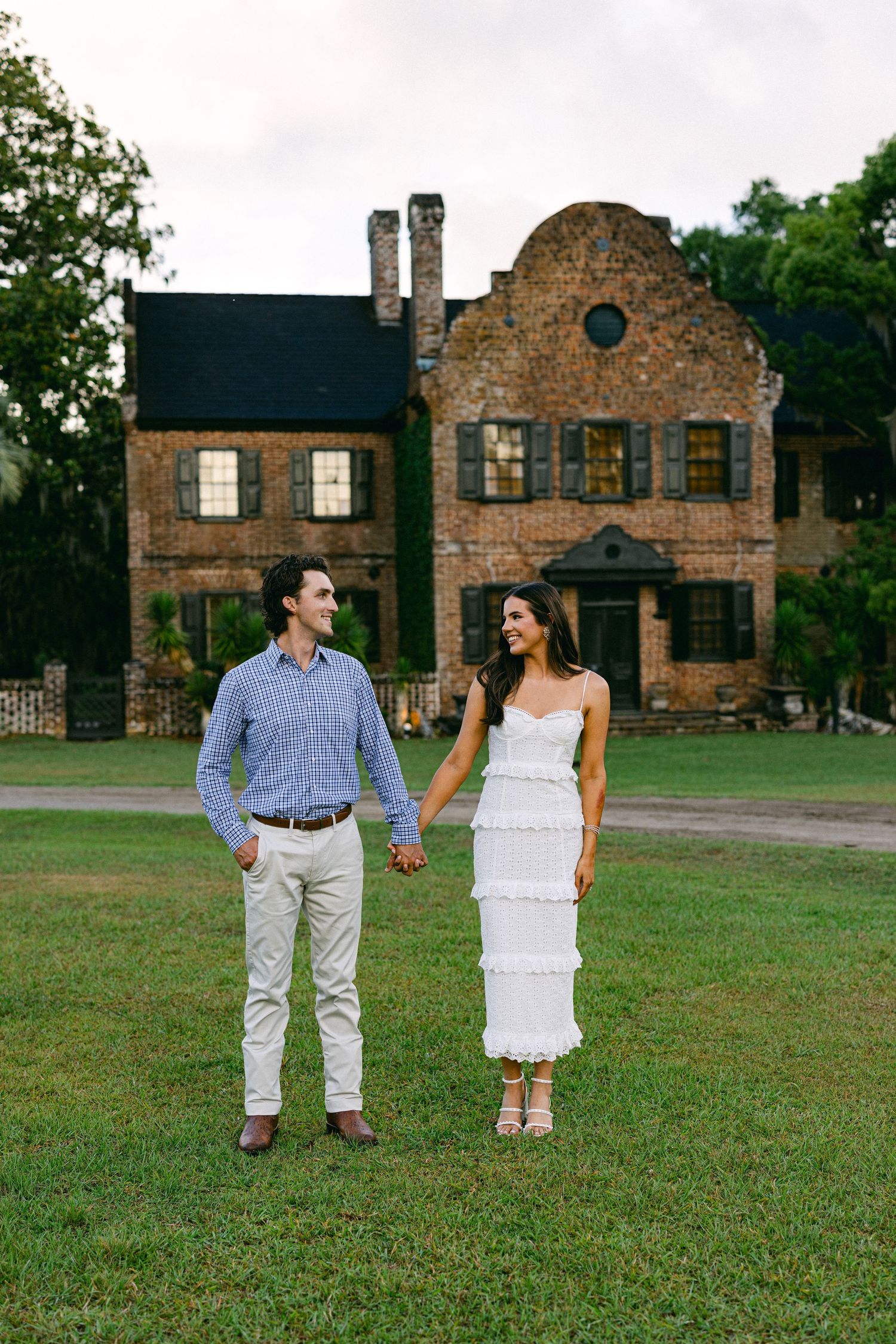 Couple holding hands in white dress and blue shirt on green lawn before historic brick mansion.