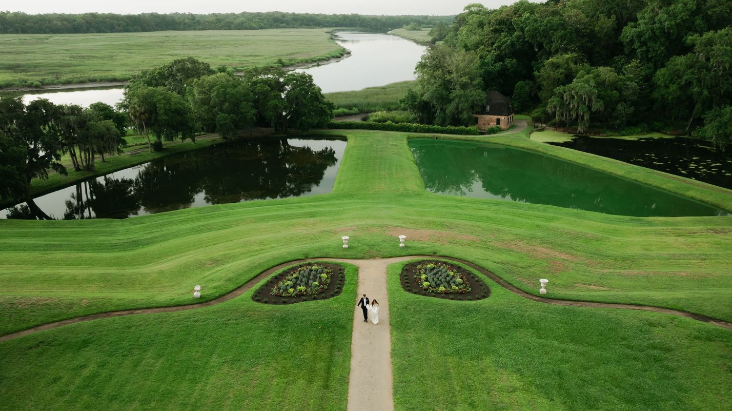 Aerial view of a lush green formal garden with symmetrical flower beds, a reflecting pond, and a river in the background.