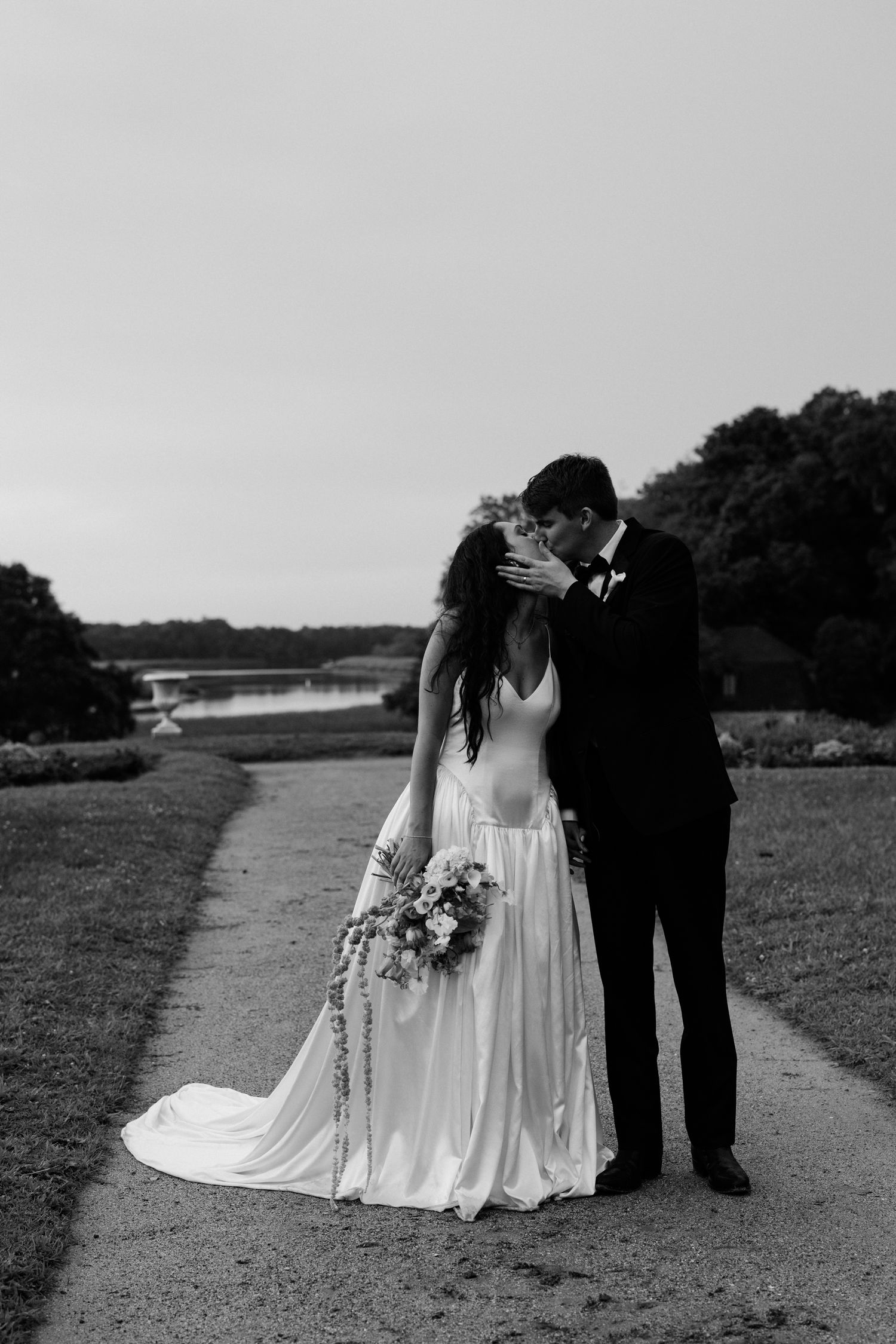 Black and white photo of a bride and groom sharing a kiss on a garden path with trees and a lake in the background.