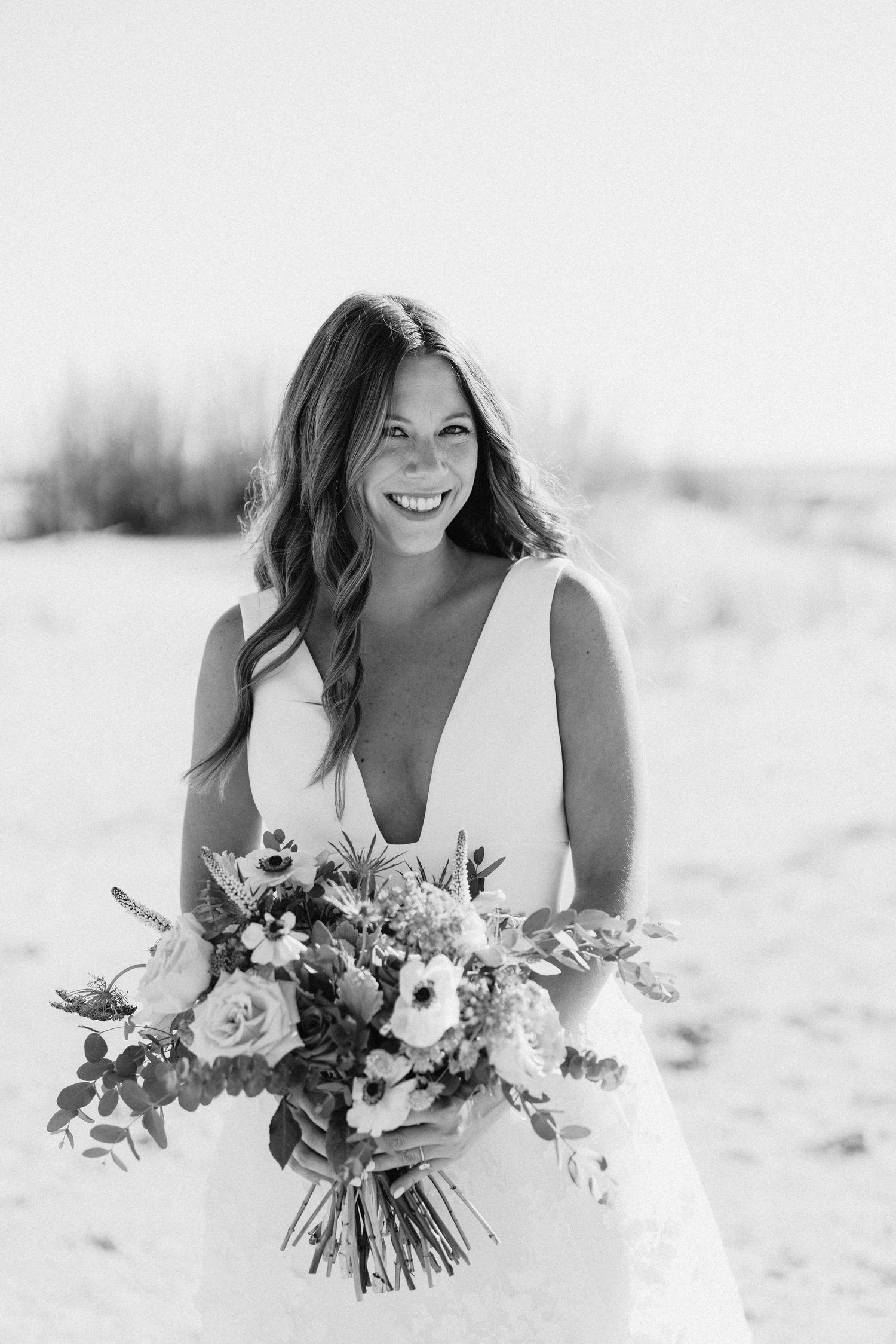 Smiling bride in a deep V-neck wedding gown holds a lush floral bouquet on a sandy beach.