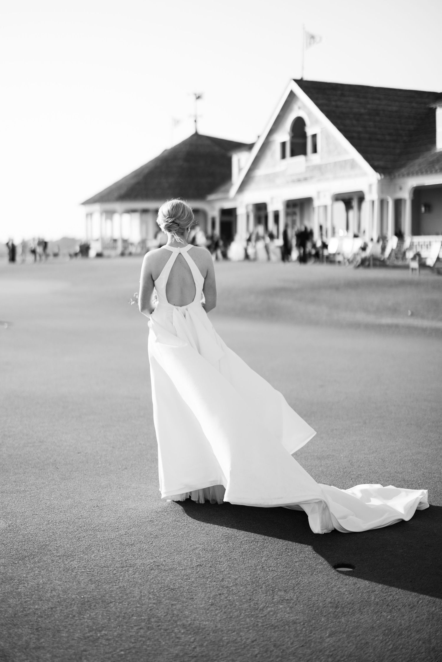 Bride in elegant backless white gown with train stands on golf course before a classic clubhouse in black and white photo.