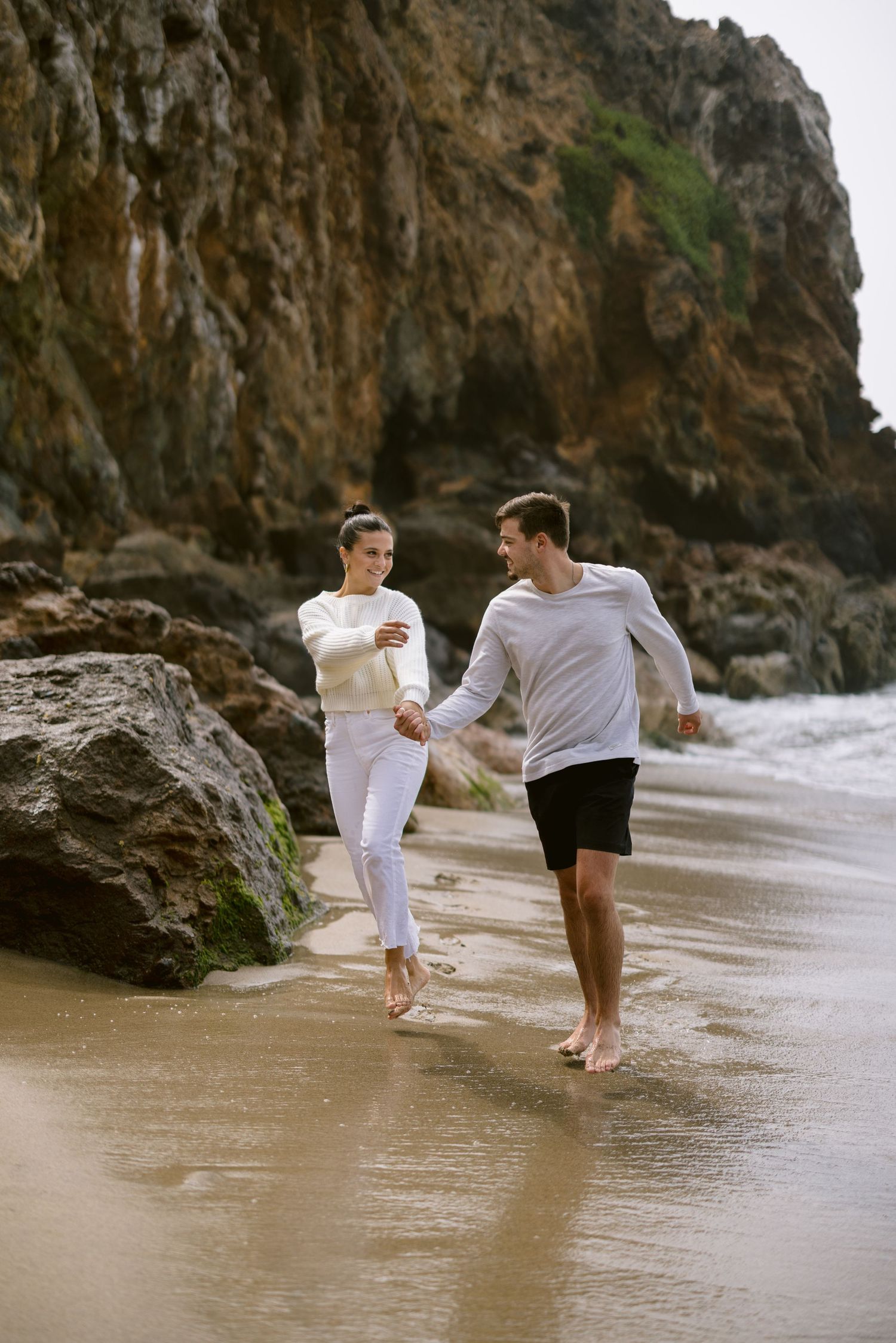 Happy couple running hand in hand along sandy beach beside large coastal rock formation.