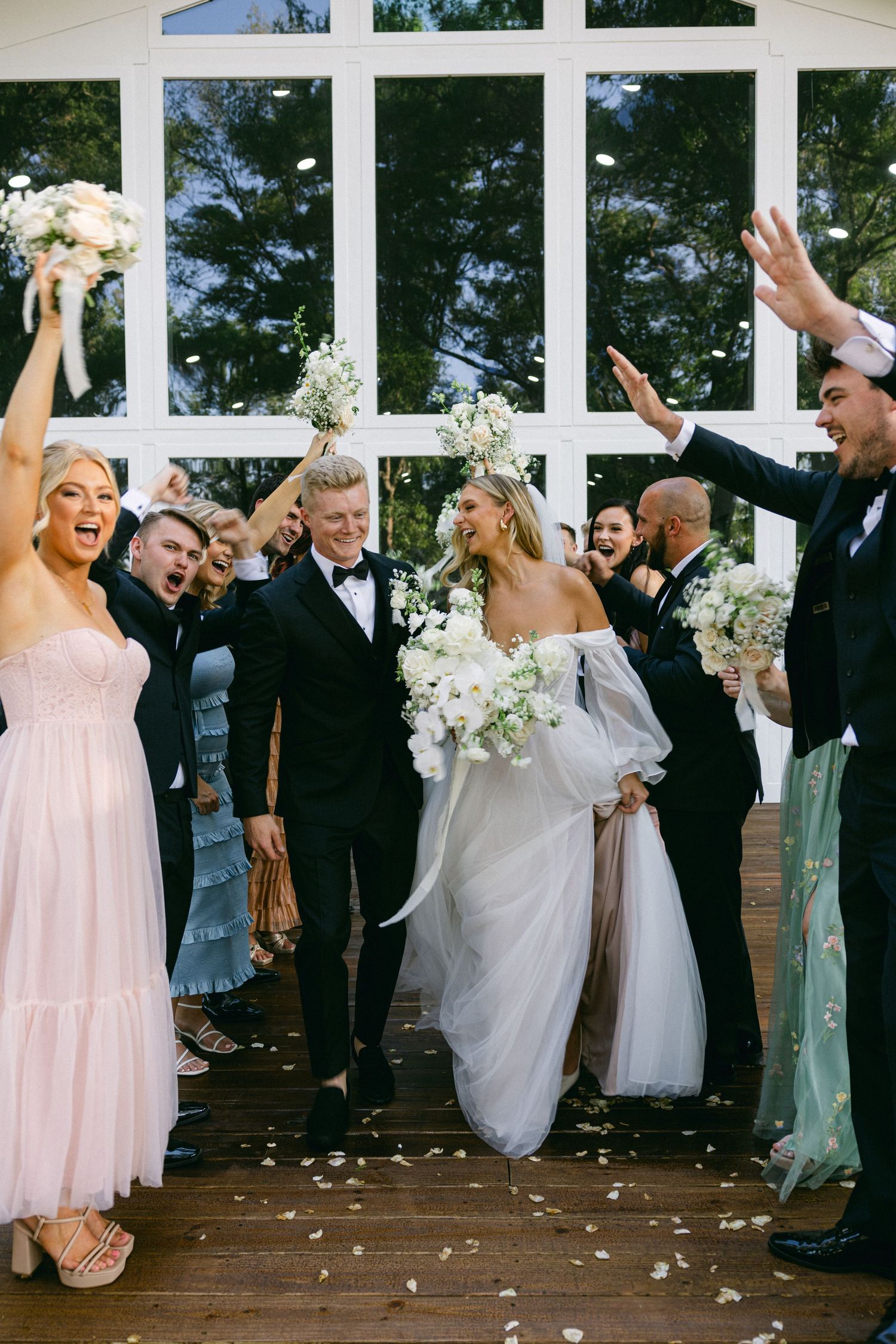 Joyful newlyweds exit chapel surrounded by cheering wedding party throwing white flower petals.