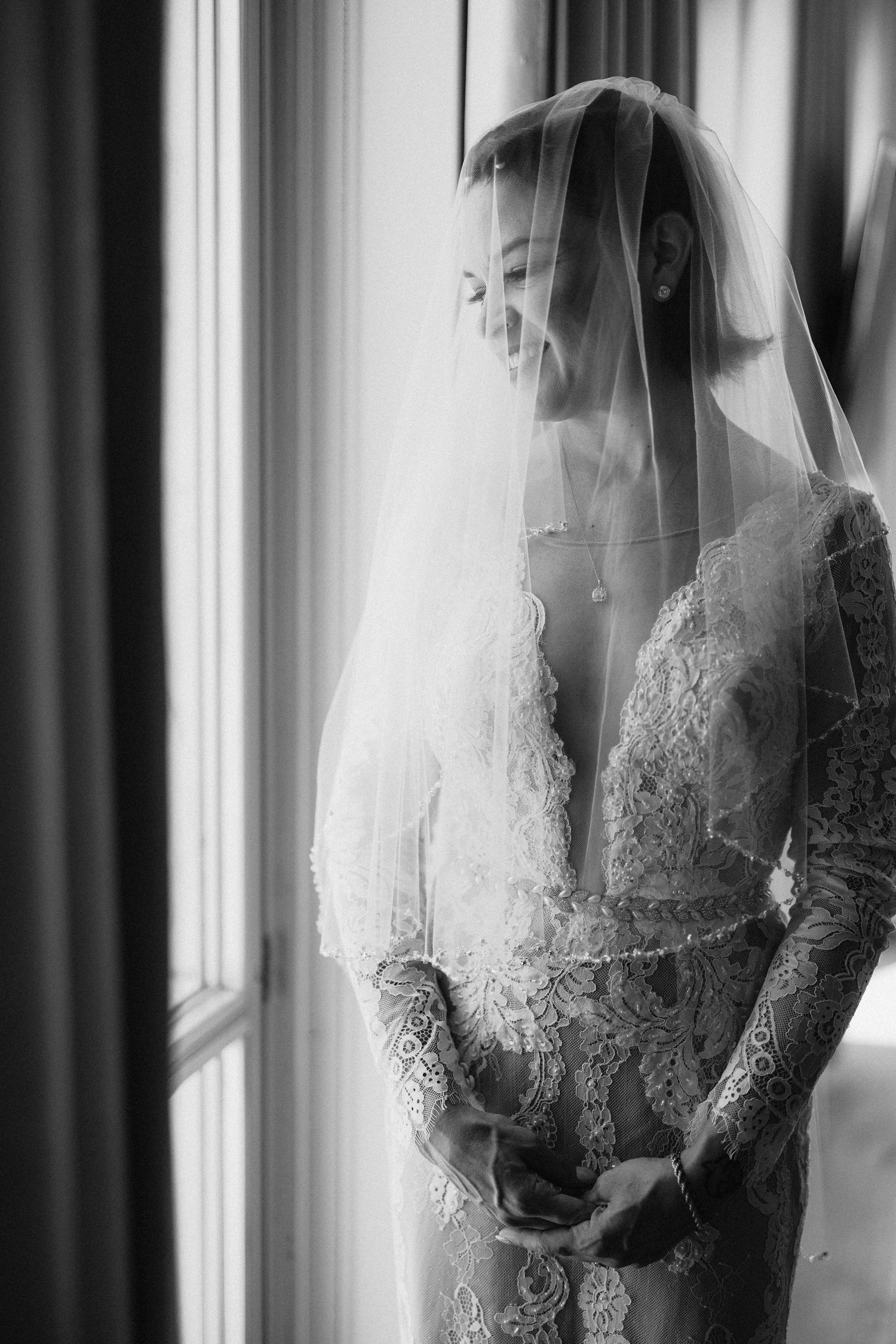 Black and white photo of a smiling bride in lace gown and veil standing by a window on her wedding day.
