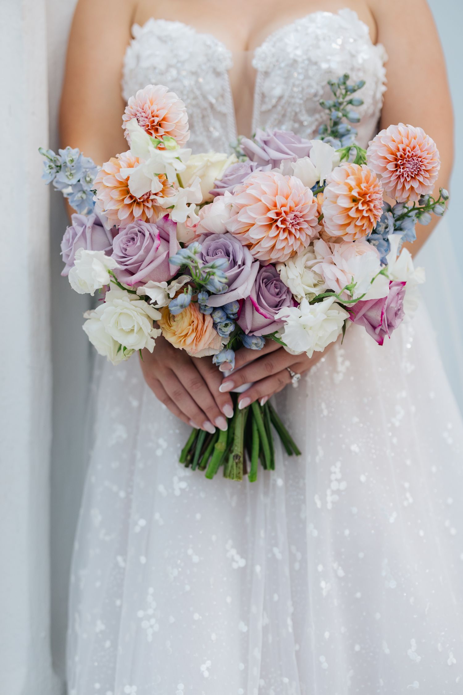 Bride holding a pastel wedding bouquet with dahlias, roses, and blue accents in a white lace gown.