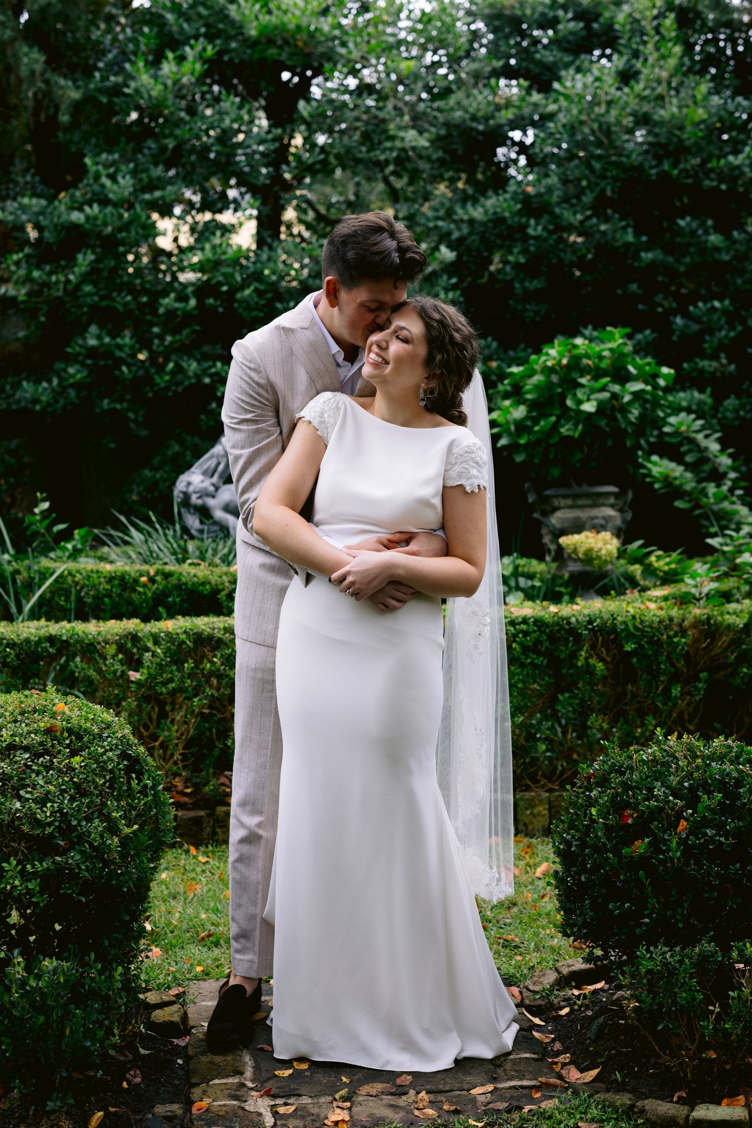Groom embracing smiling bride in white gown and veil in a lush garden setting on their wedding day.