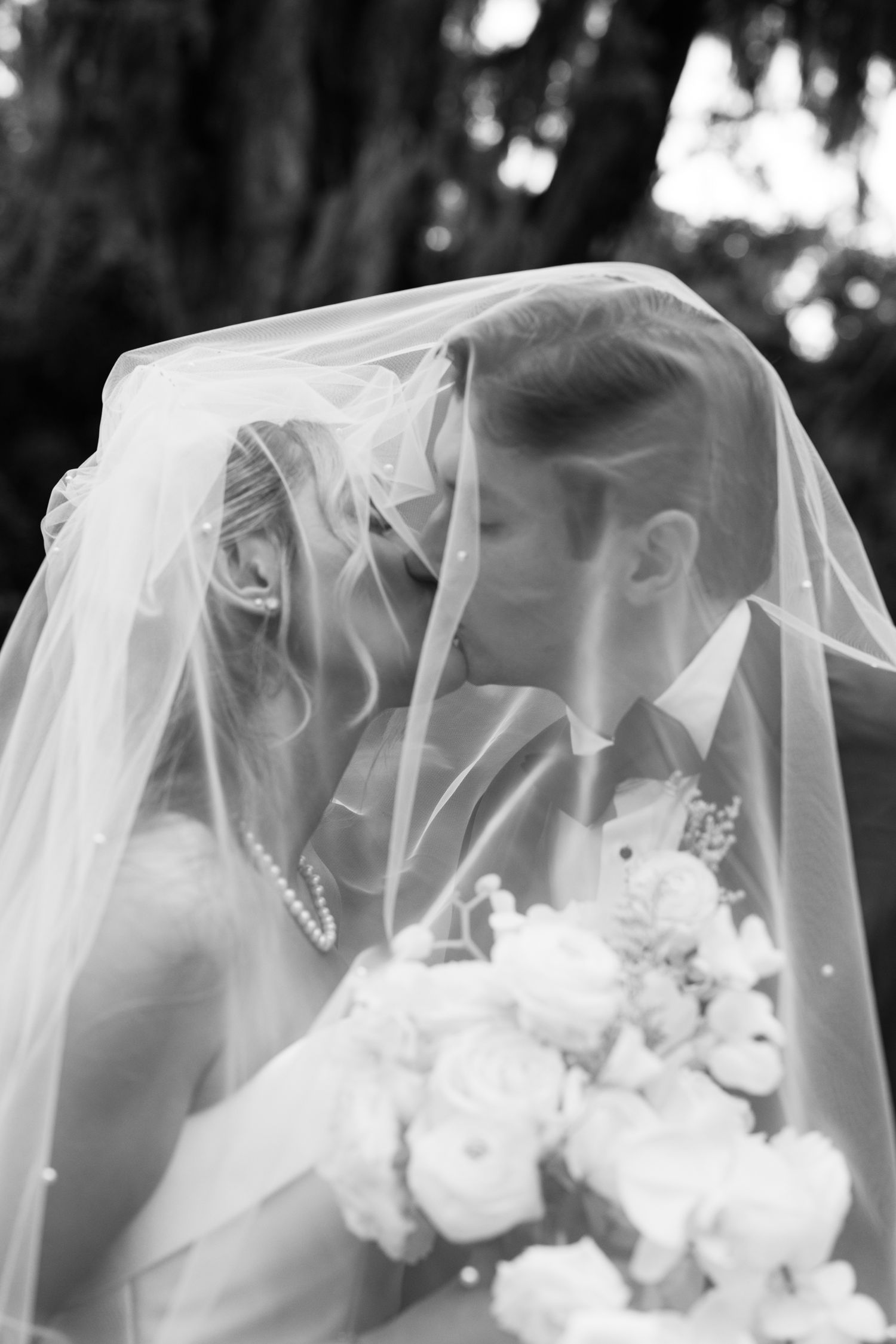 Black and white photo of bride and groom kissing beneath flowing veil holding white floral bouquet.