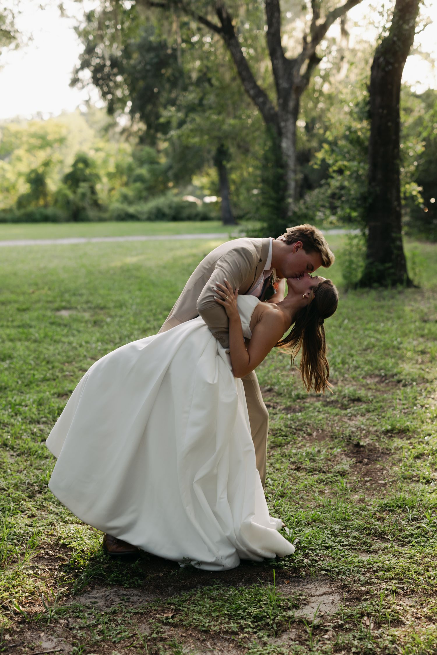 Groom dipping bride for a romantic kiss outdoors on green grass, surrounded by lush trees in a garden setting.