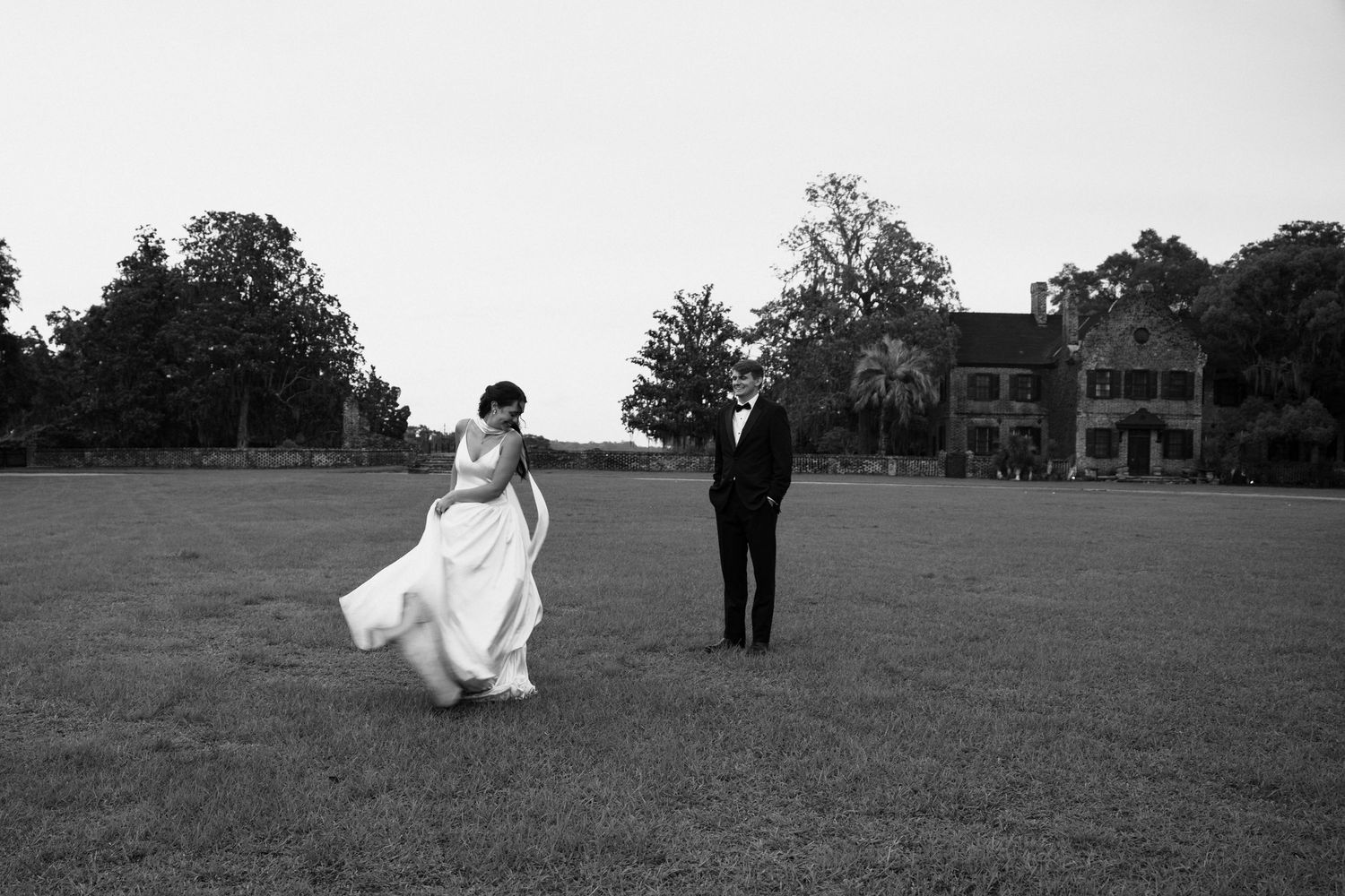 Black and white photo of bride and groom walking apart on expansive lawn with historic manor in background.