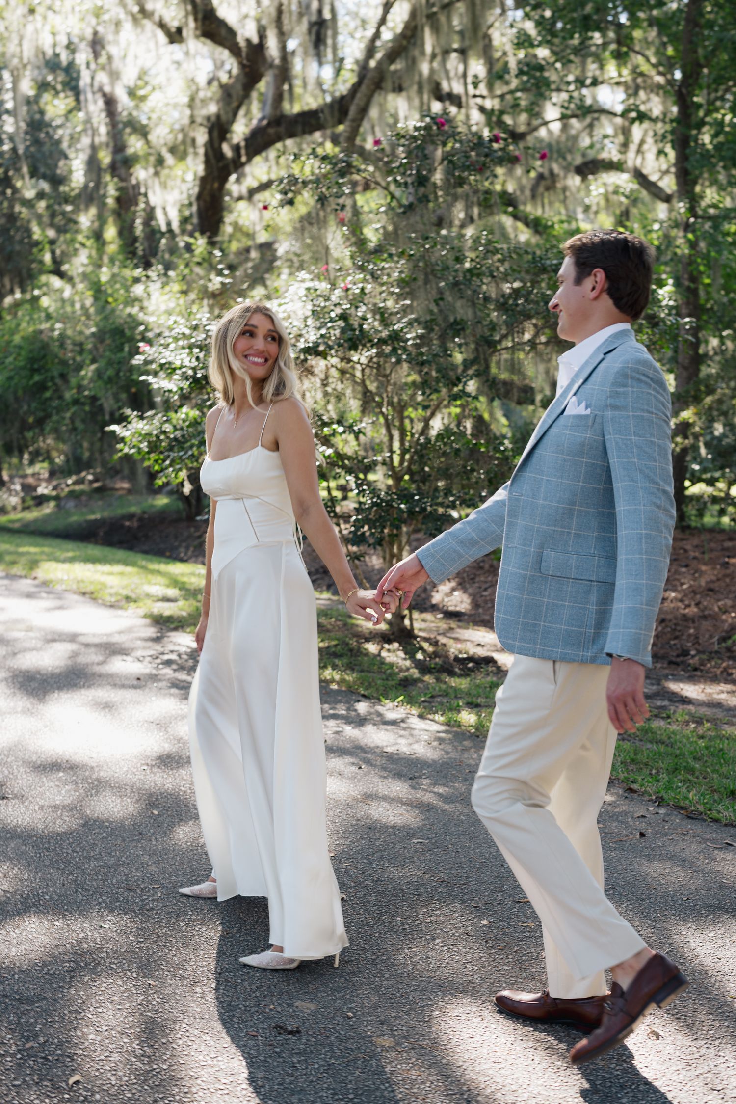 Bride in white jumpsuit and groom in light blue blazer hold hands walking outdoors under Spanish moss trees.