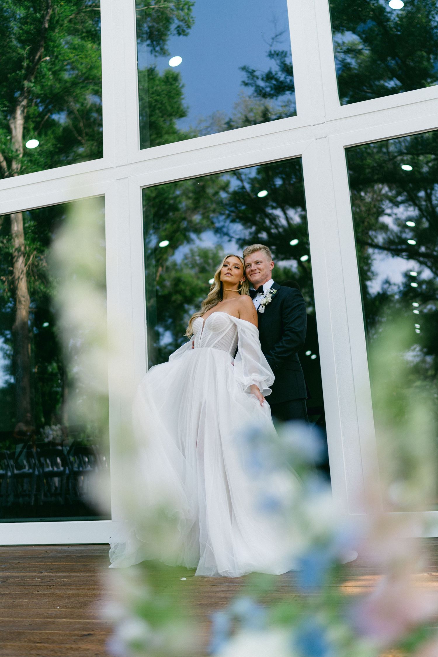 Bride in flowing white off-shoulder gown stands with groom in black tuxedo before large glass windows at elegant wedding venue.