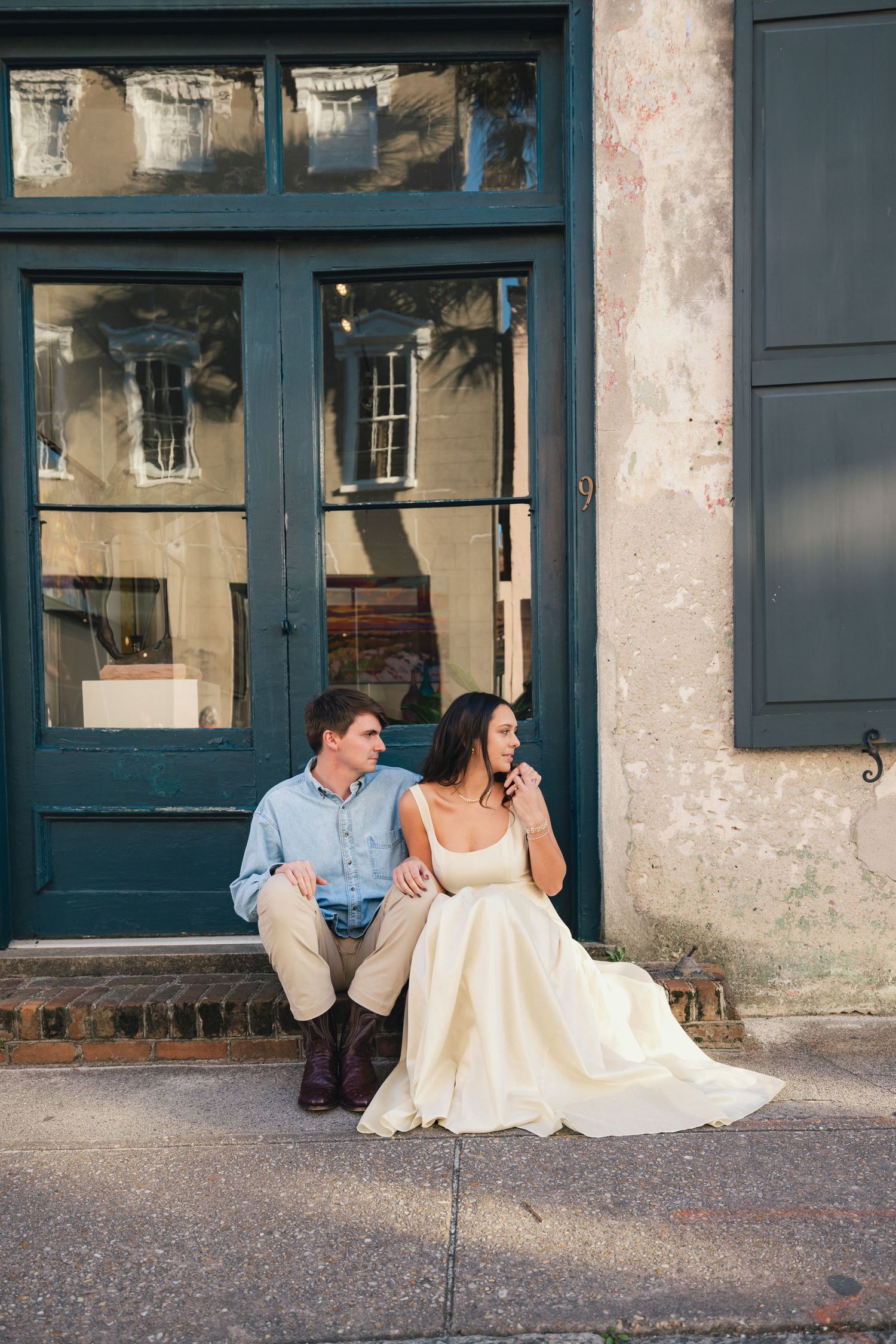 A couple in wedding attire sits on a sidewalk in front of a teal door on a charming historic street.