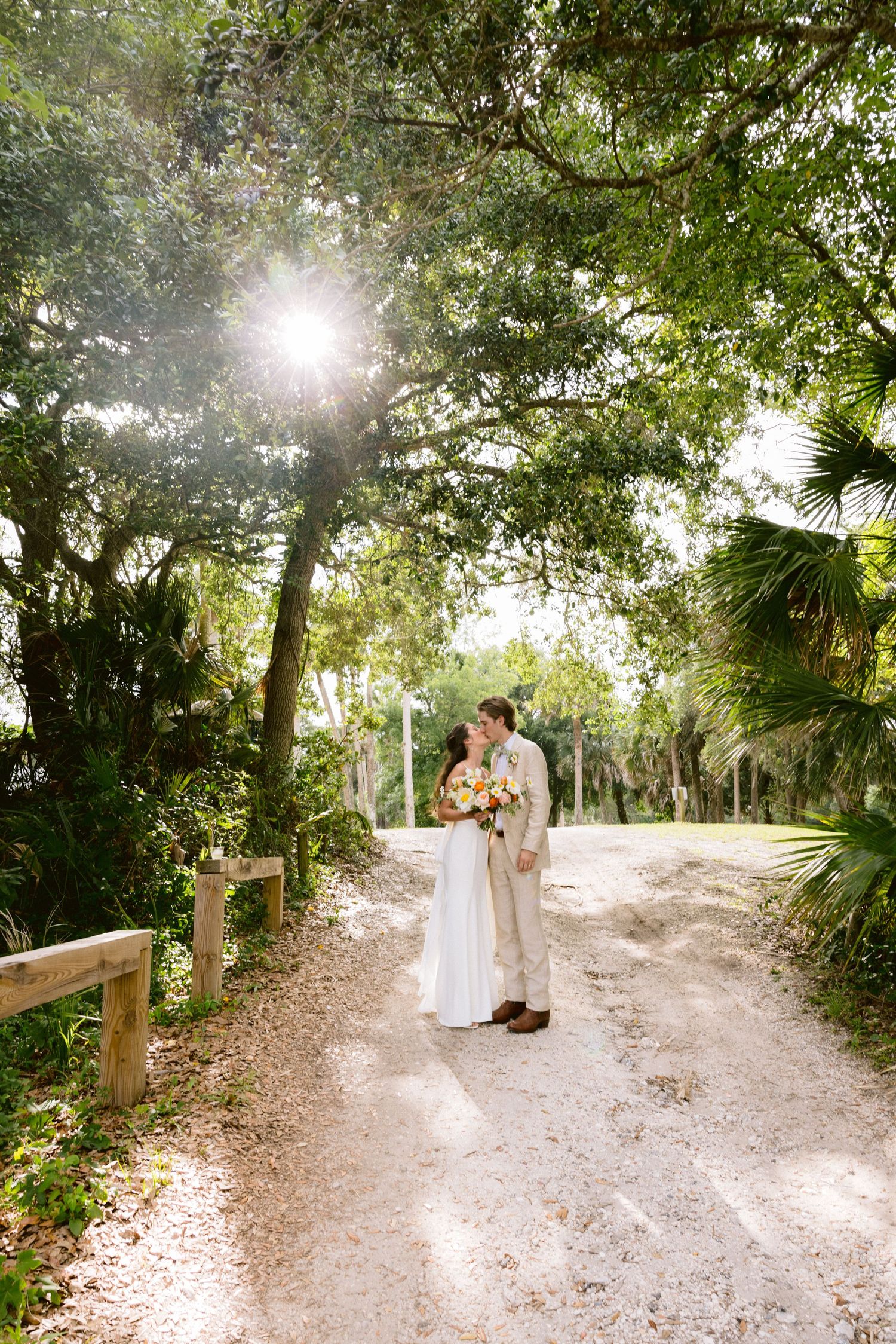 Bride and groom kissing on a sunlit sandy path surrounded by lush green trees at an outdoor wedding.