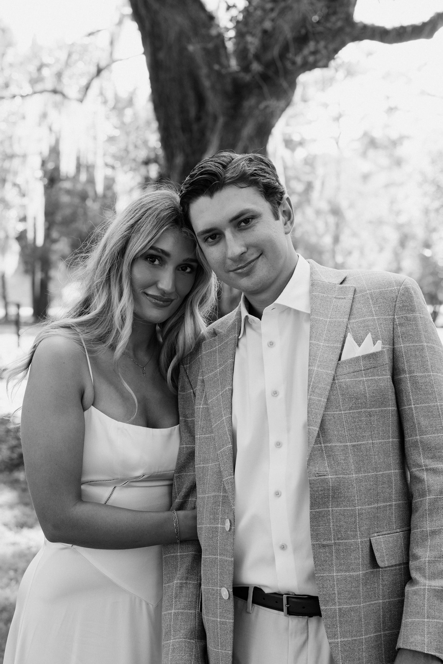 Black and white photo of a smiling couple posing outdoors, woman in white dress and man in plaid suit jacket.