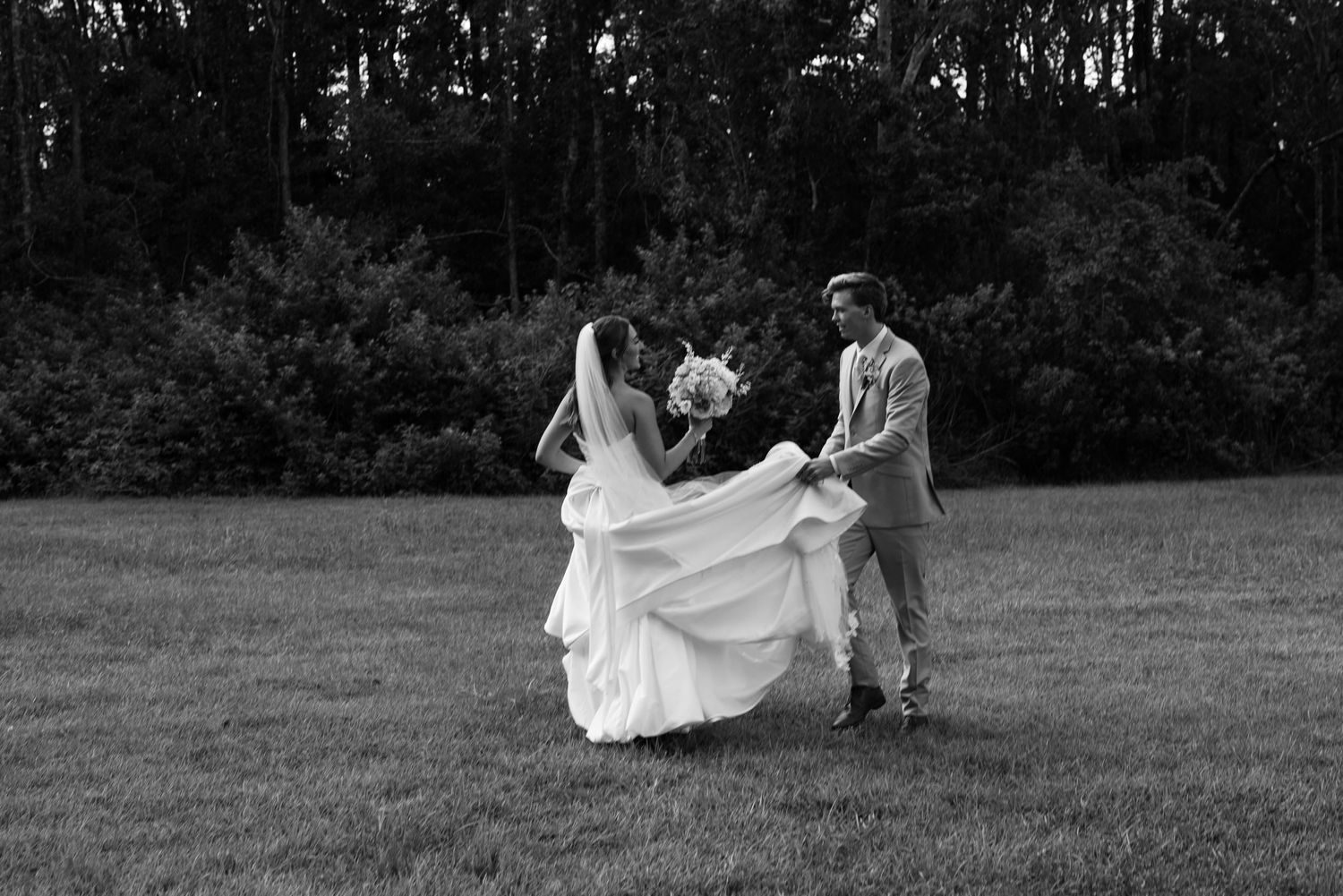 Black and white photo of bride and groom dancing playfully in an open grassy field with tree backdrop.