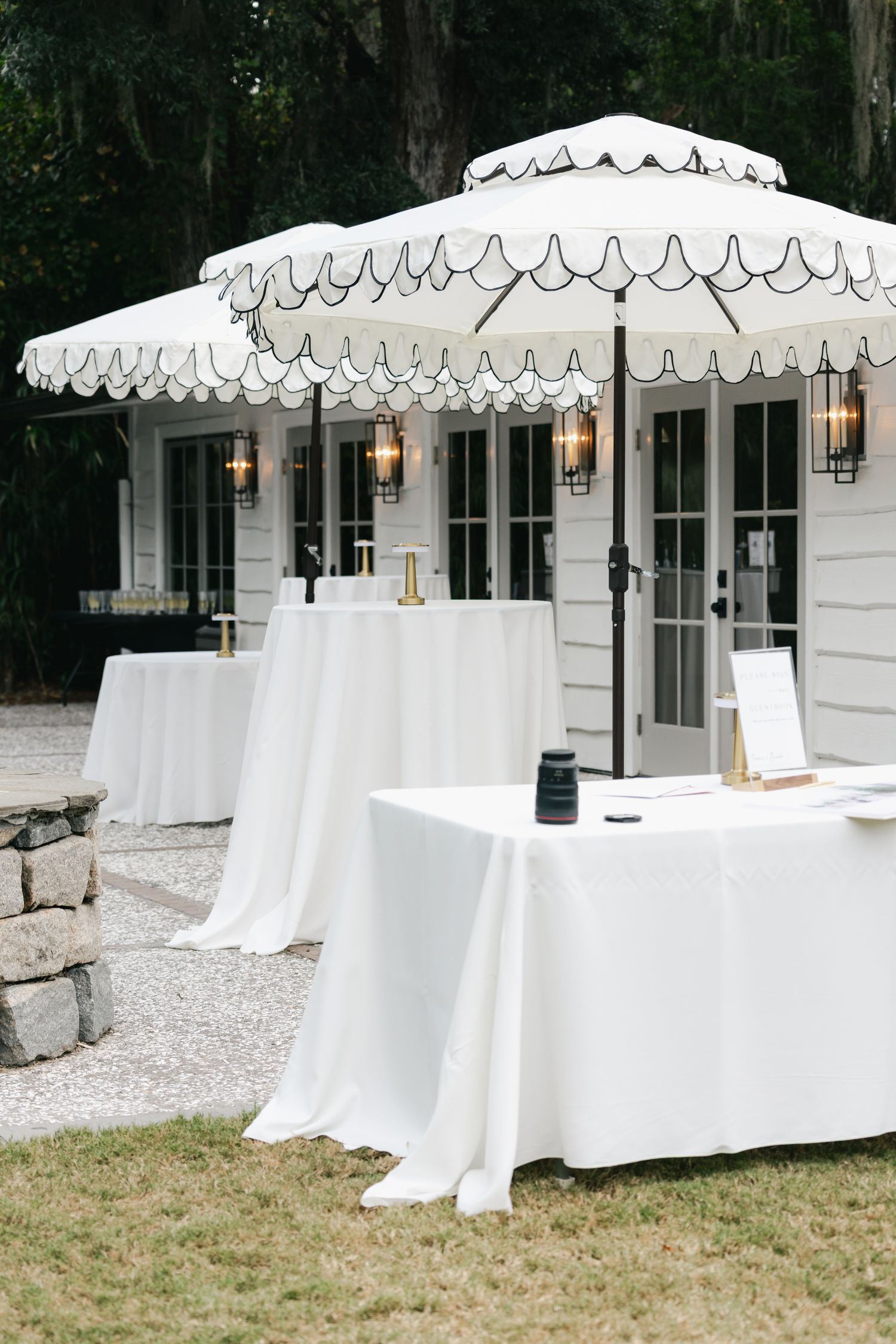Outdoor wedding cocktail hour setup with white scalloped umbrellas, draped tables, and lantern-lit white building.
