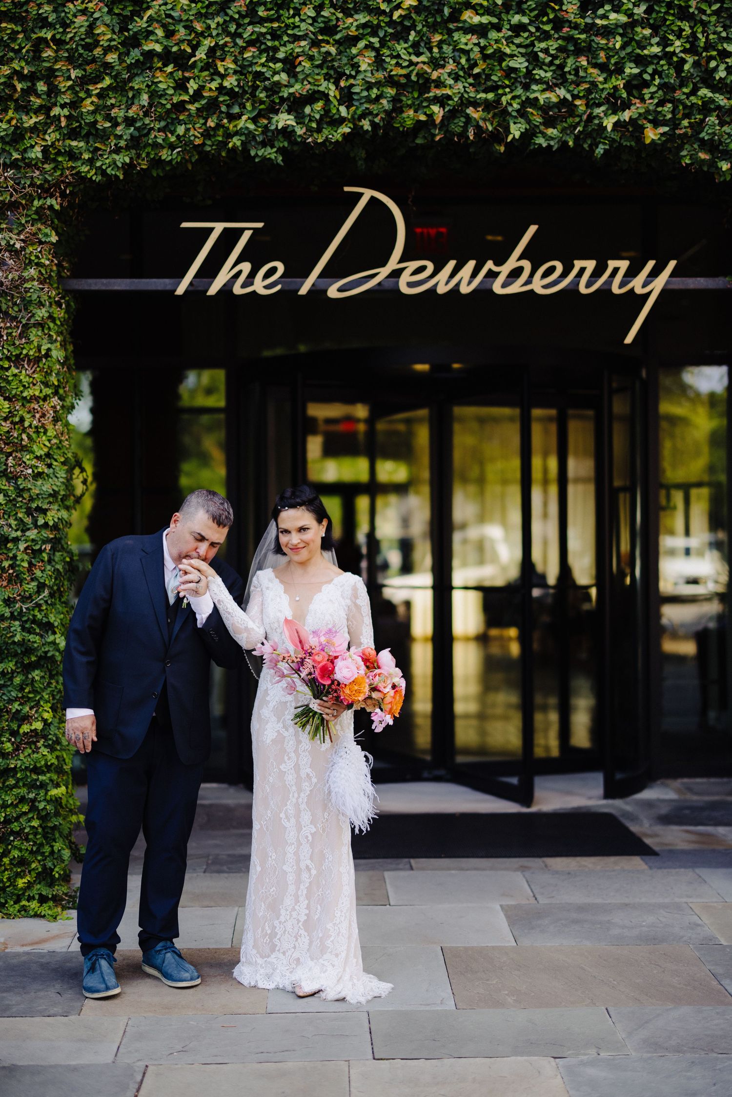 Bride and groom pose outside The Dewberry hotel, bride holding colorful bouquet with feathers in lace wedding gown.