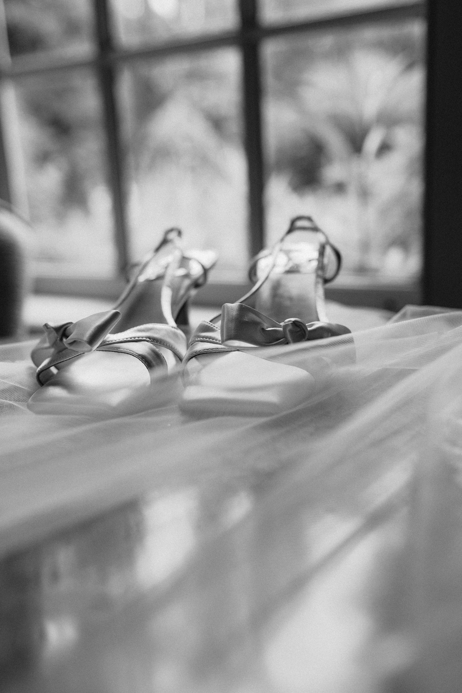 Black and white photo of elegant bridal sandals resting on a veil near a sunlit window.
