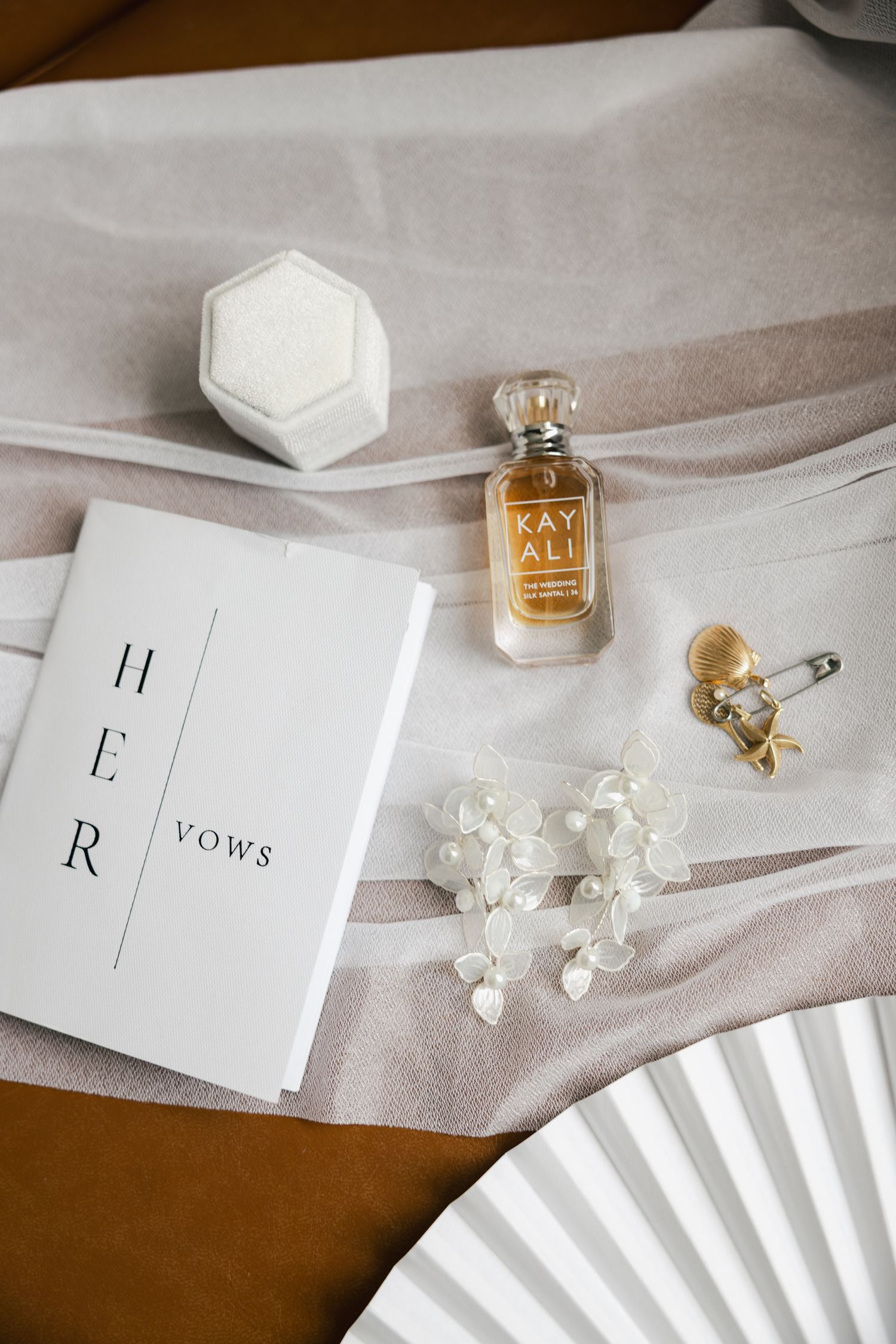 Elegant wedding flat lay with vow book, Kayali perfume, ring box, floral earrings, and fan on neutral fabric.
