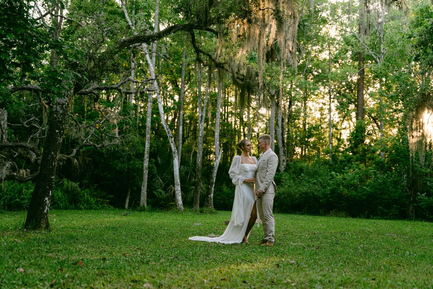 Bride and groom dancing on lush green lawn surrounded by moss-draped trees at golden hour.
