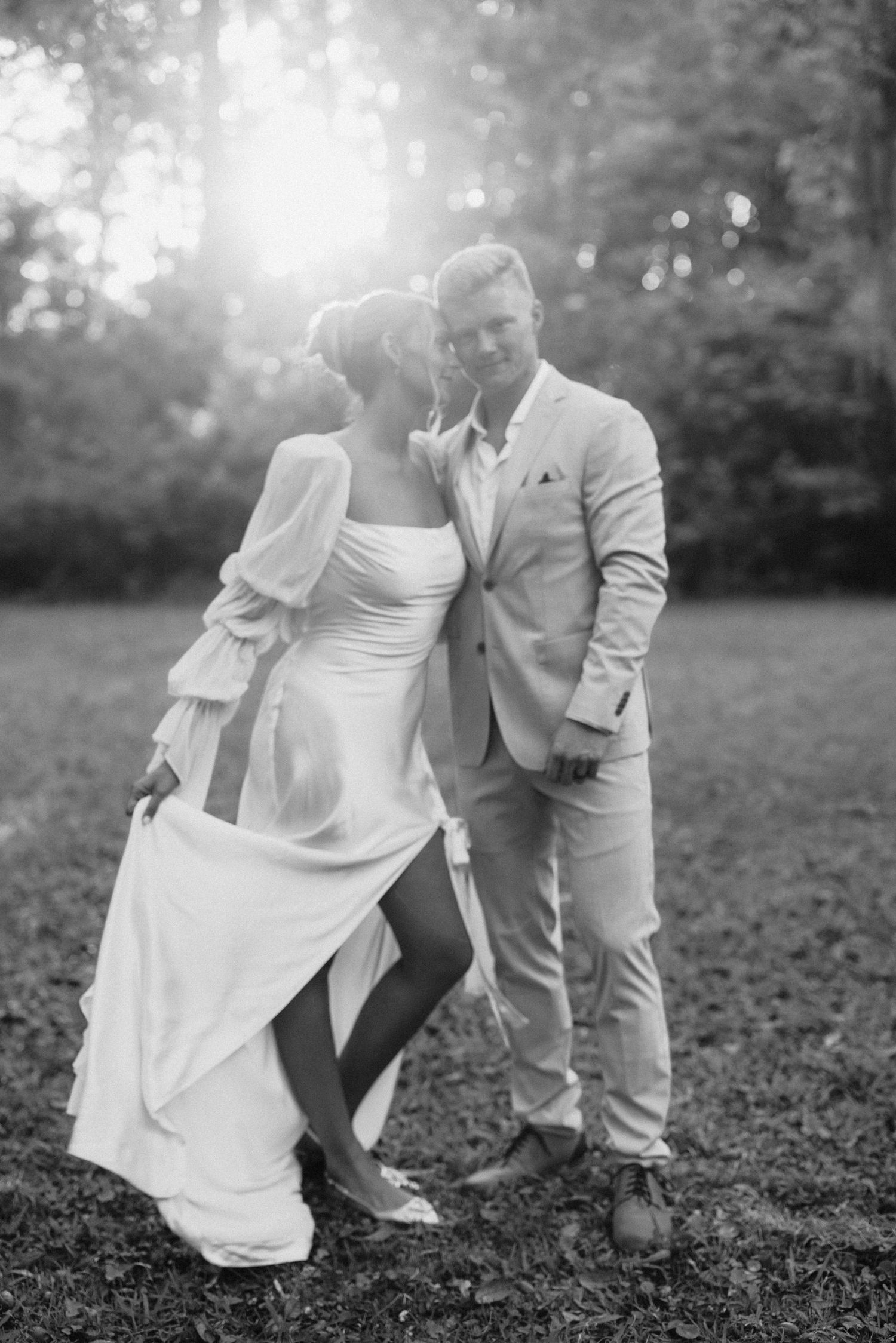 Bride and groom embracing in sunlit garden, bride in flowing white gown with sheer sleeves, groom in beige suit.
