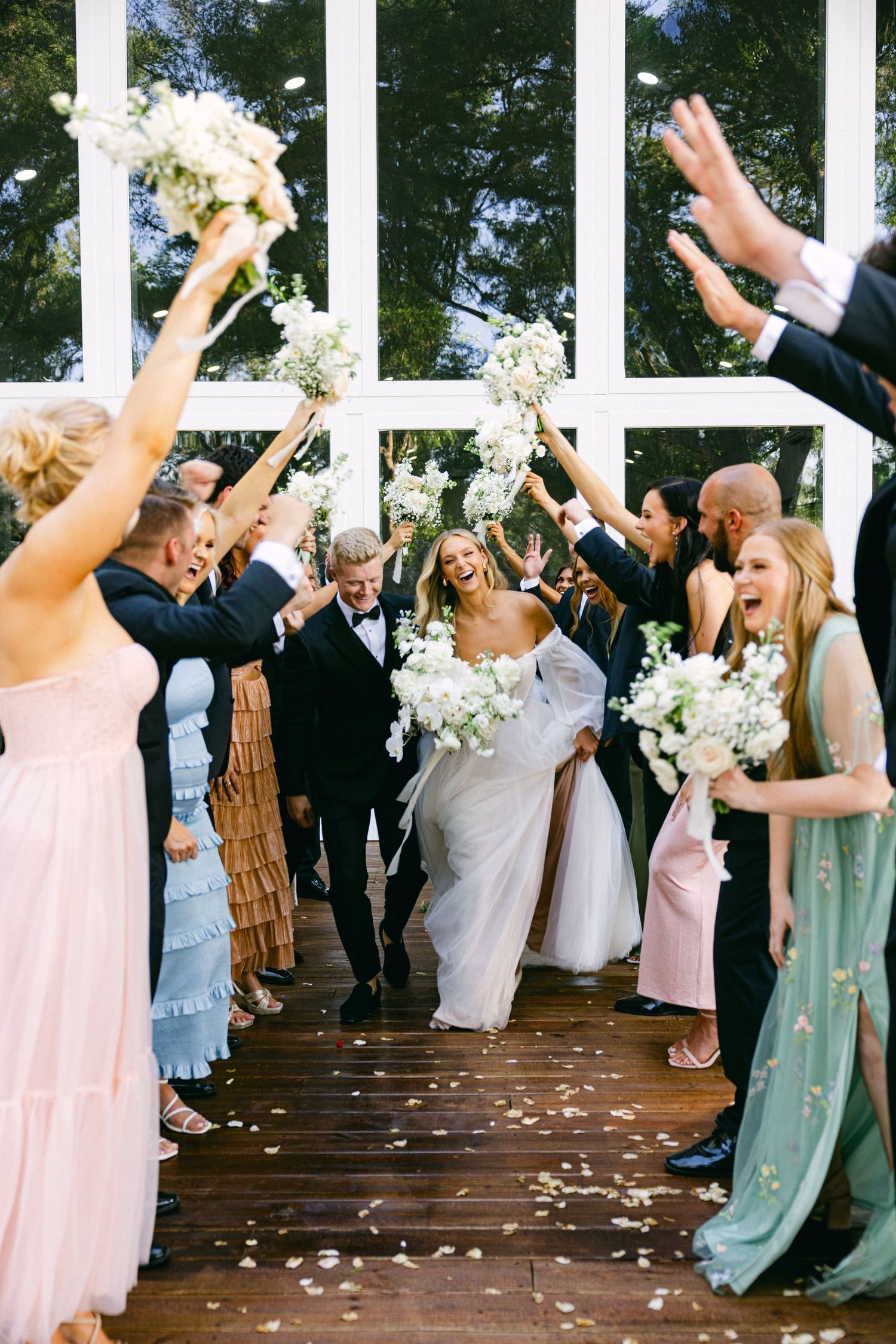 Bride and groom walk through cheering wedding party holding white floral bouquets on wooden deck.
