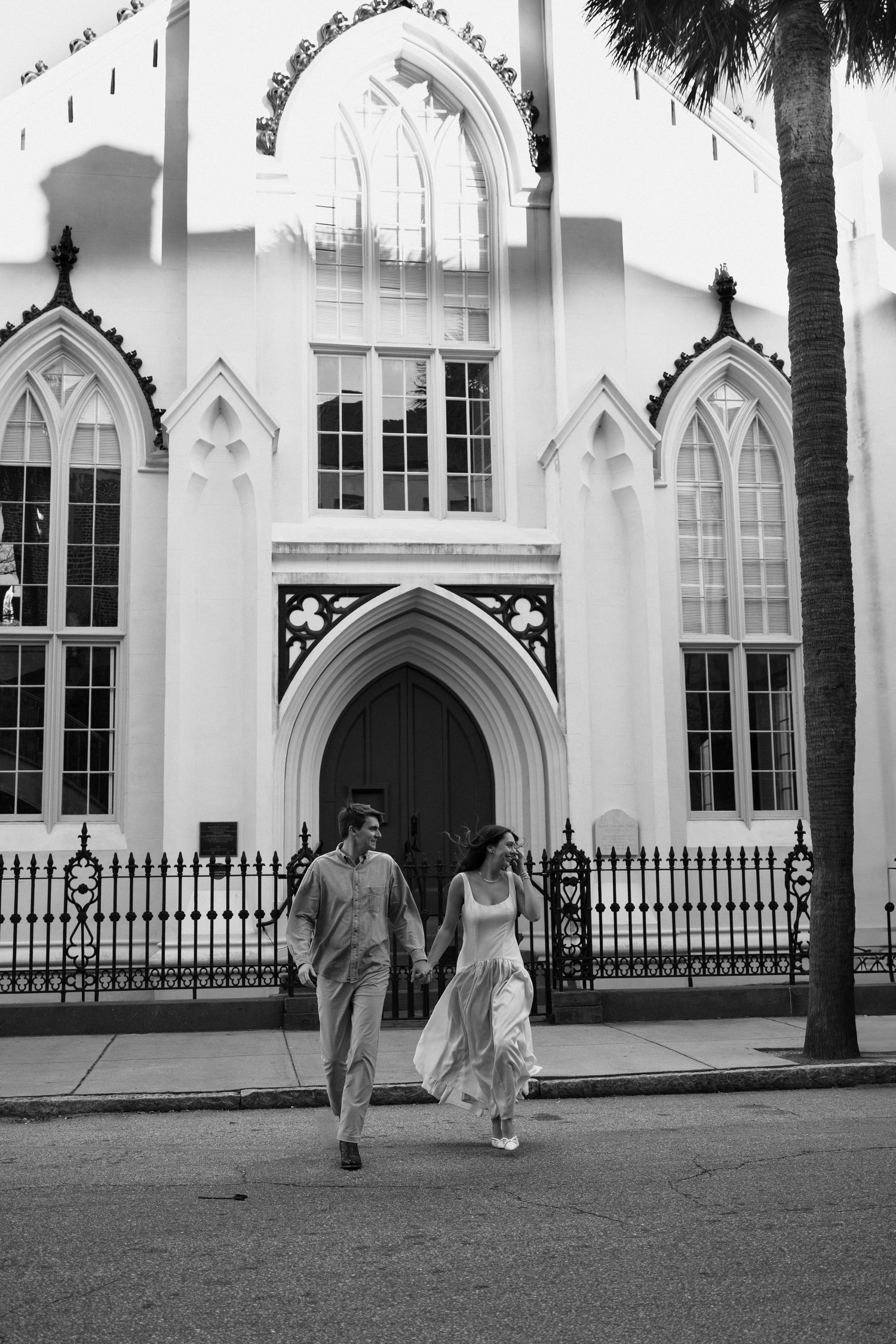 Newlywed couple holding hands walking in front of a Gothic church in black and white wedding photo.