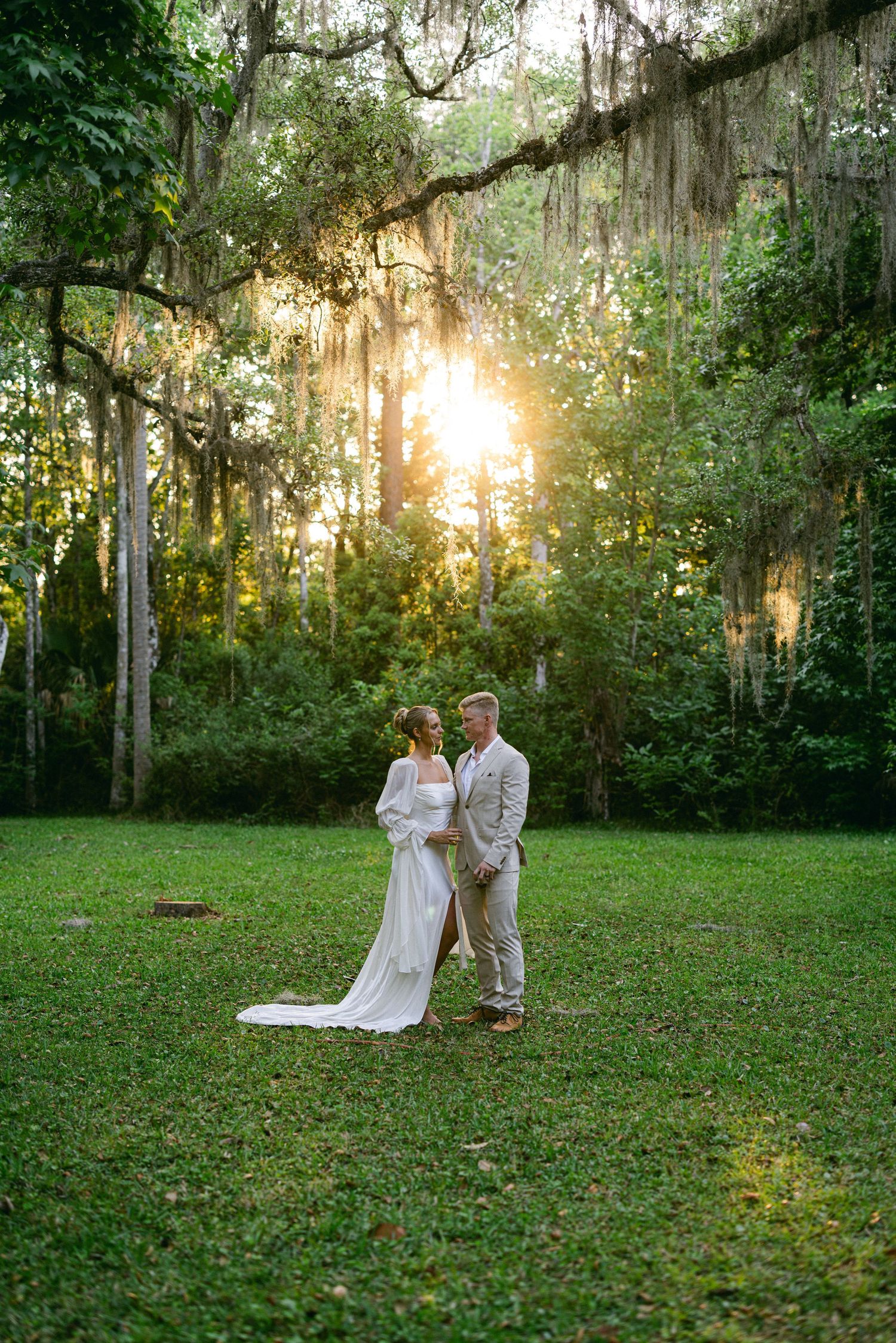 Bride and groom standing together in golden sunset light beneath sweeping moss-draped trees.
