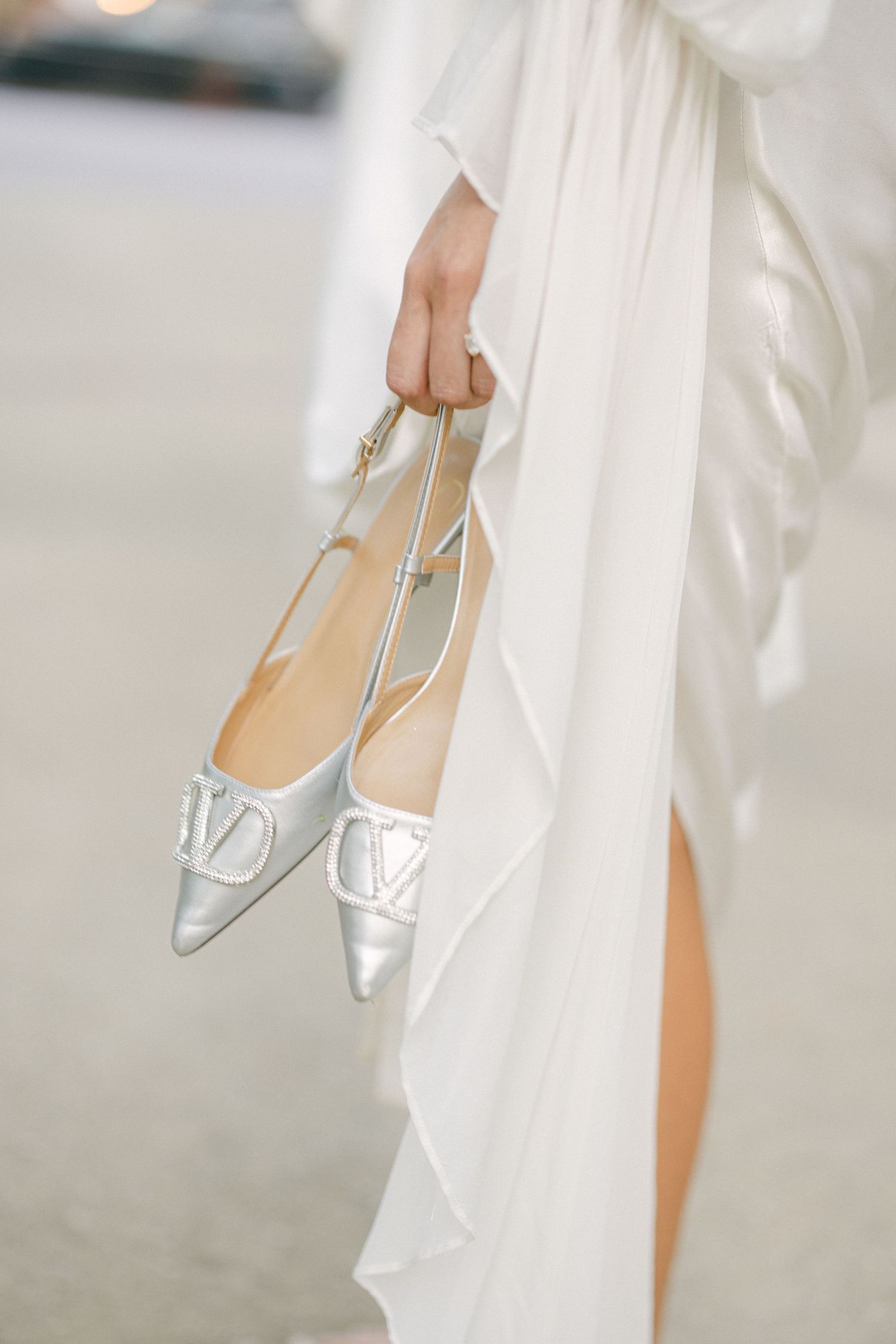 Bride holding silver Valentino pointed-toe flats against flowing white wedding dress with dramatic sleeves.