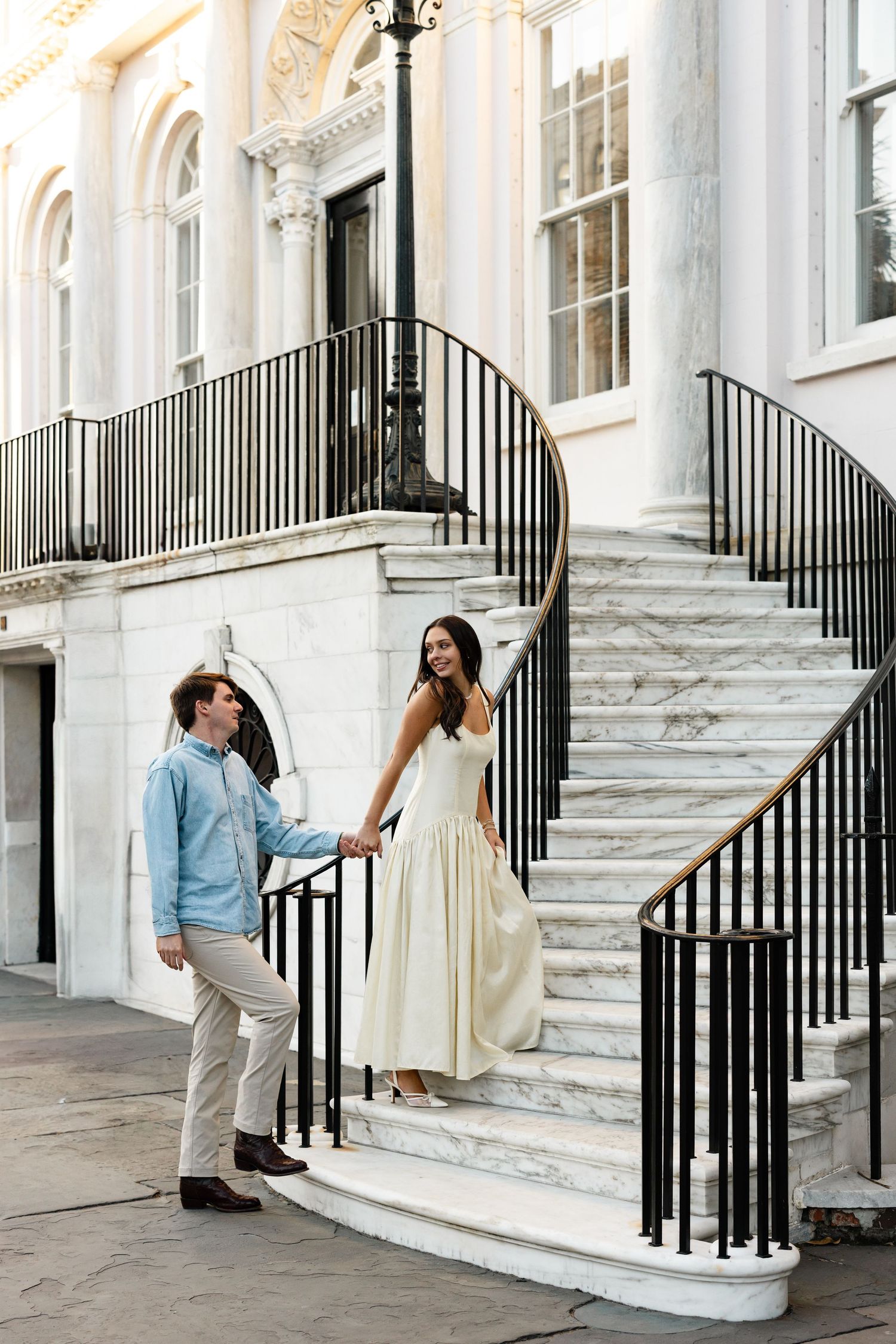 Couple holding hands on elegant marble staircase, woman in white dress, man in blue shirt, classic building backdrop.