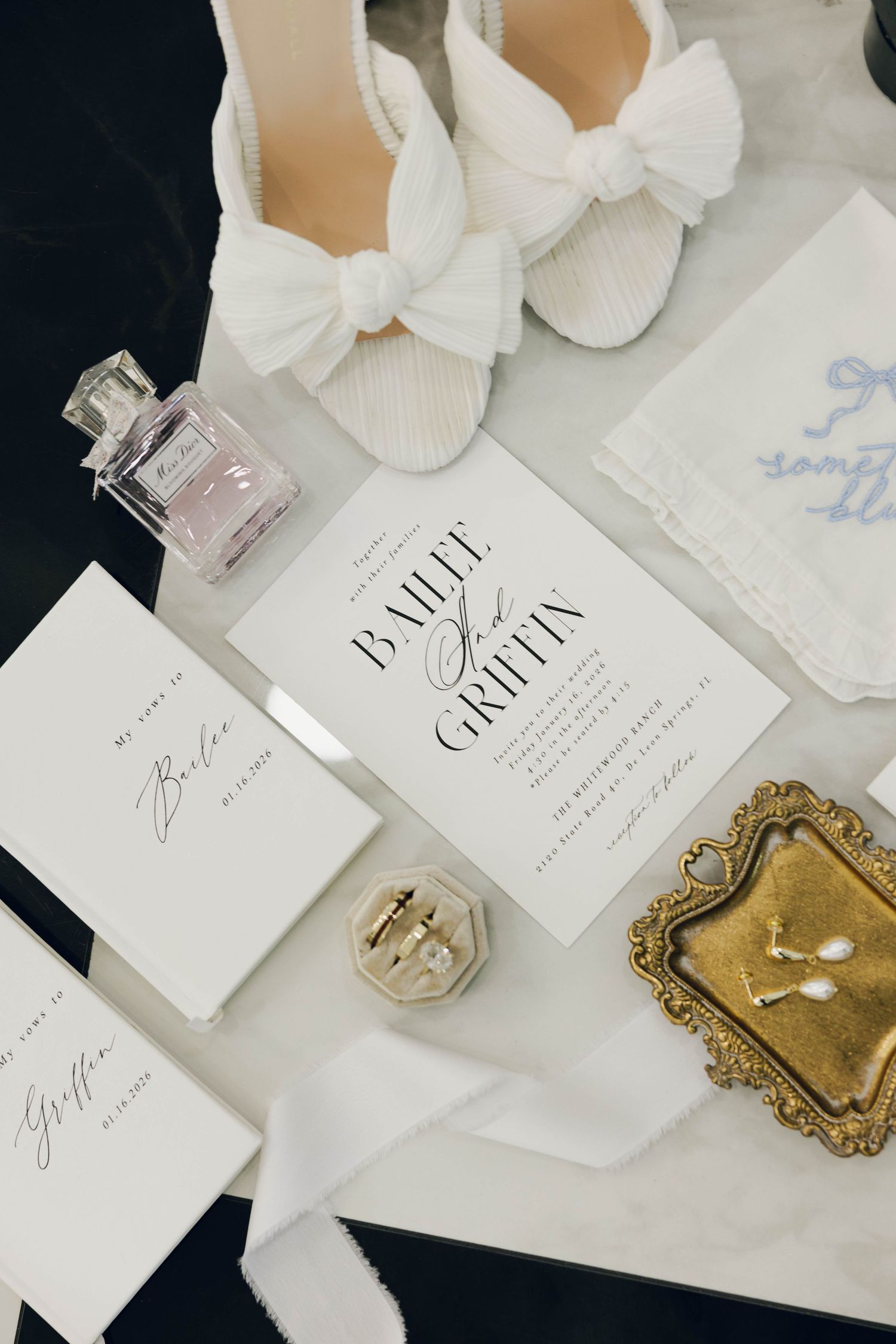 Elegant wedding flat lay with white bow heels, floral perfume, gold ring dish, invitation suite, and bridal accessories on marble.