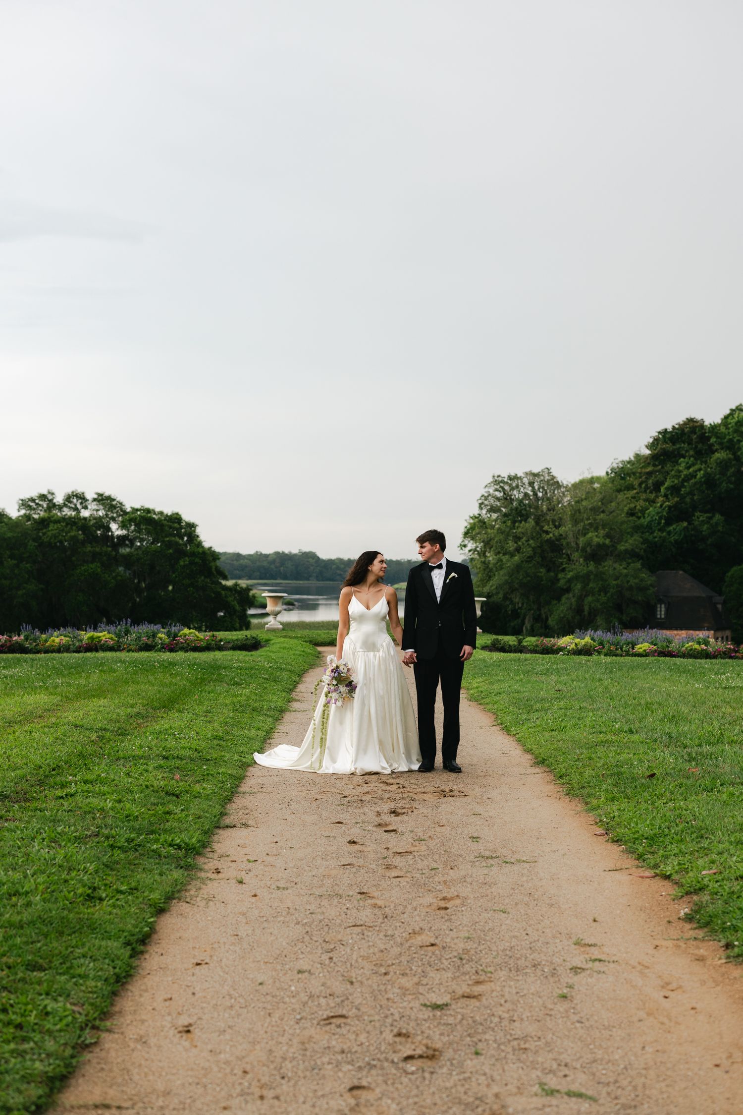 Bride and groom walk hand-in-hand down a scenic garden path, bride in white gown holding bouquet, groom in black tuxedo.