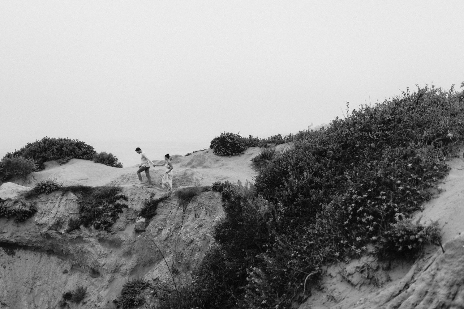 Couple standing on a sandy coastal bluff with wild vegetation in black and white photography.