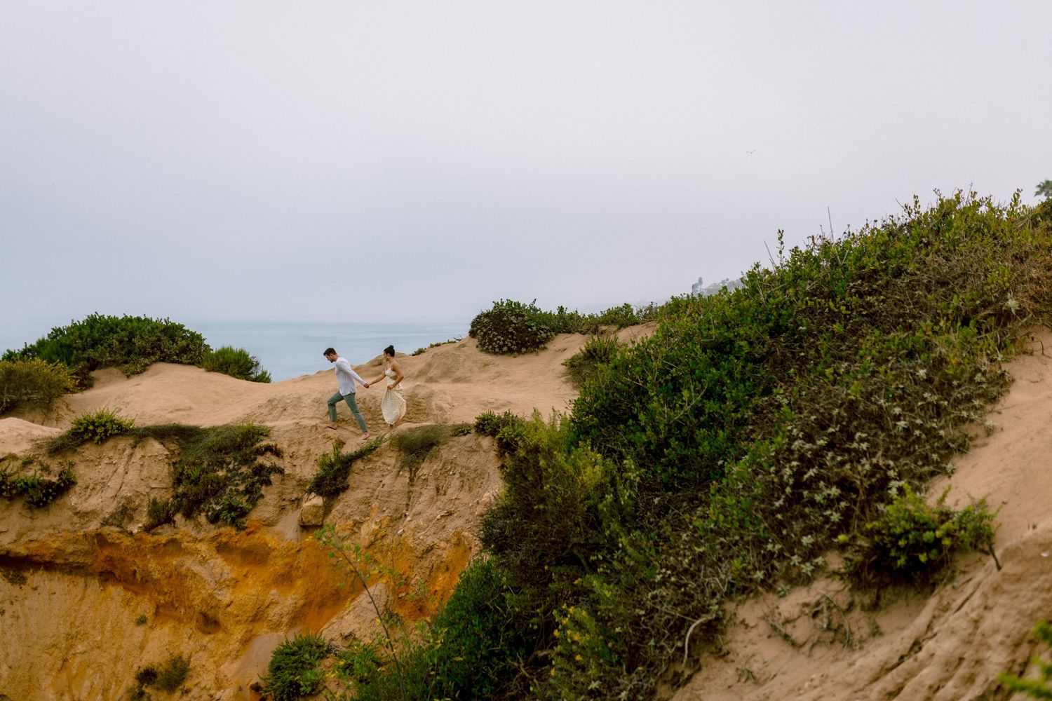 Couple holding hands while walking along sandy coastal bluff with ocean view.