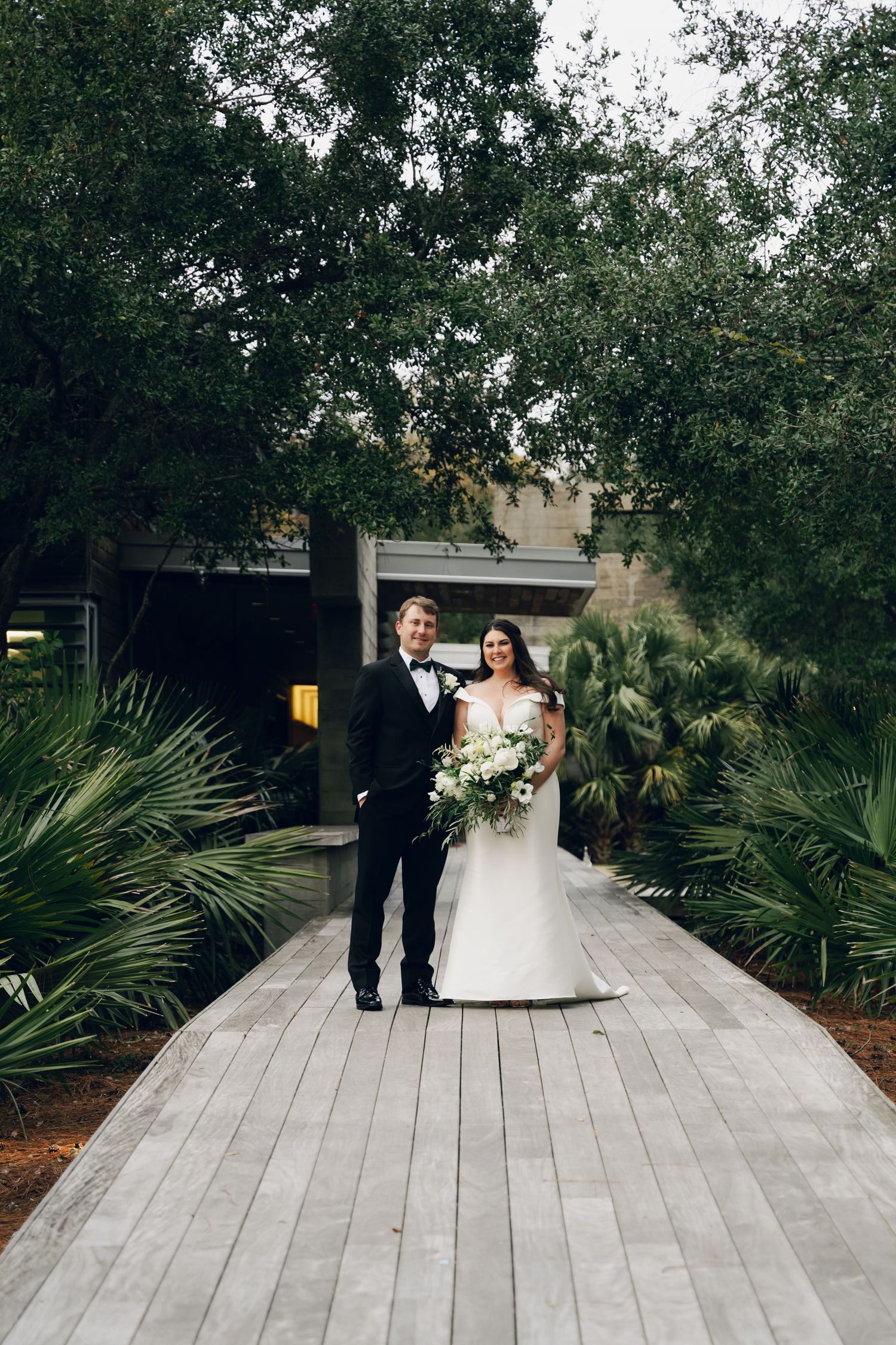 Bride and groom posing on wooden walkway surrounded by lush greenery, bride holding white bouquet.