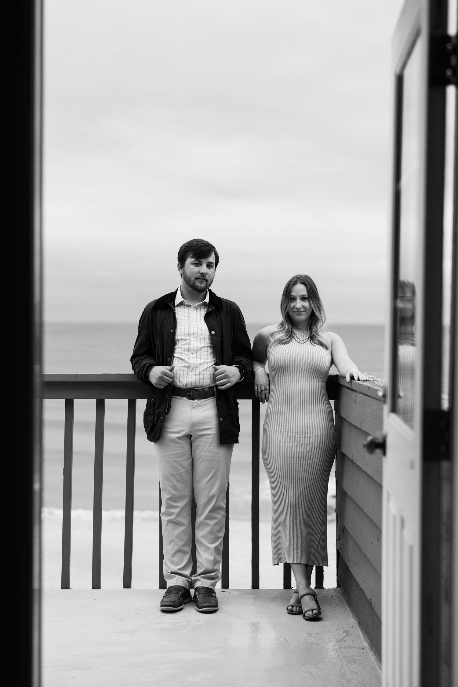 Black and white photo of a couple standing on a beach balcony with ocean views behind them.