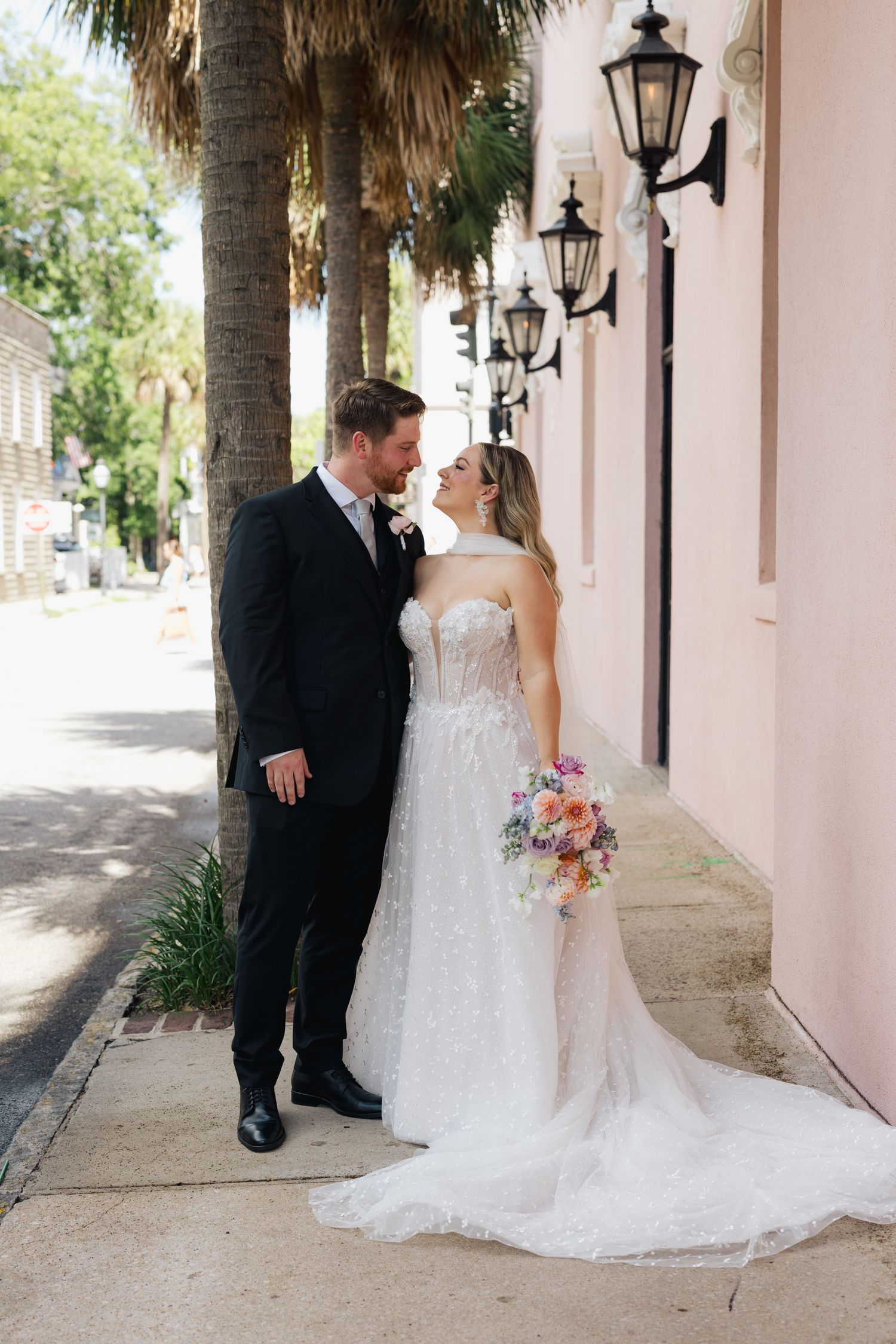 Bride and groom share a romantic moment on a Charleston sidewalk, bride holding colorful bouquet in elegant strapless gown.