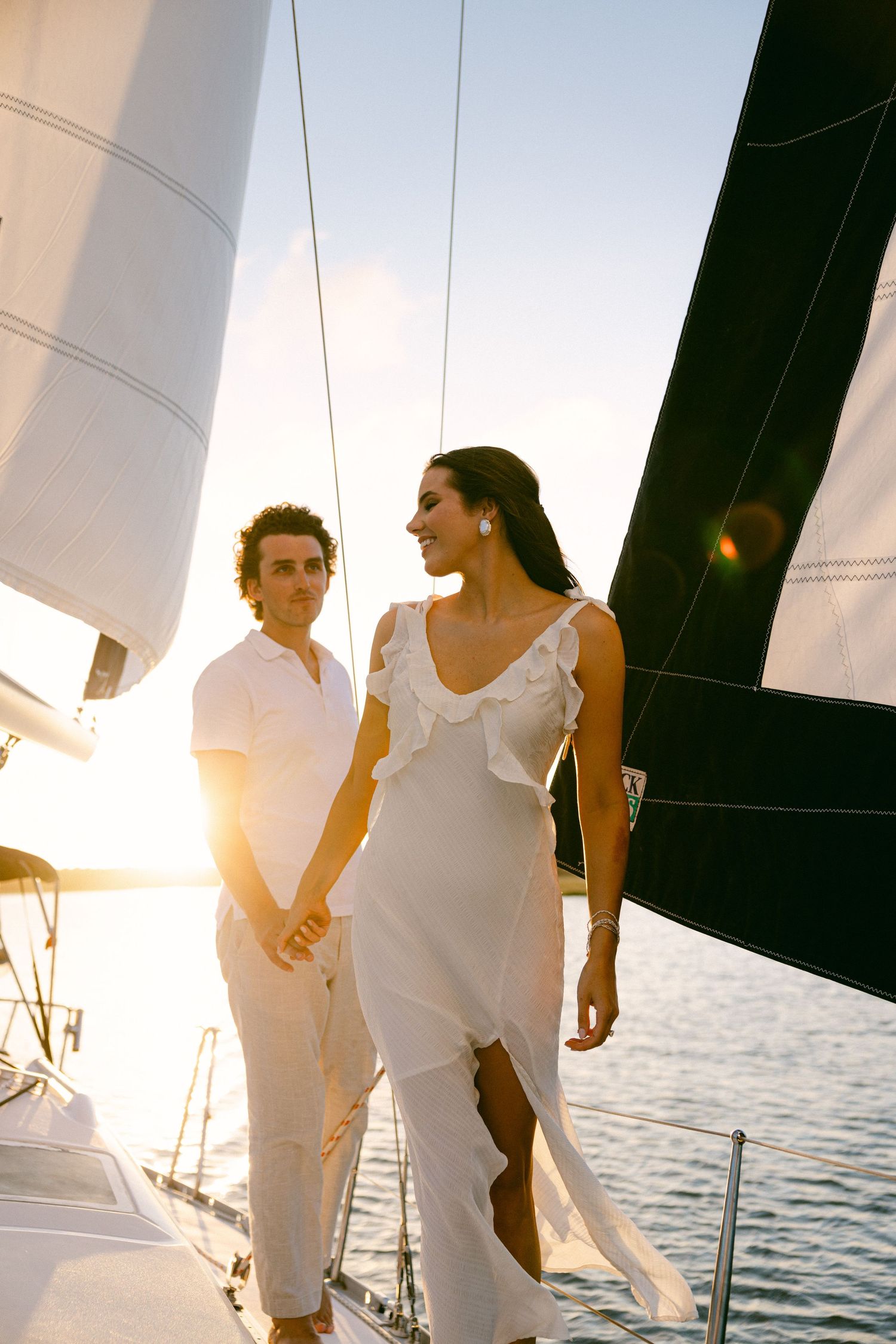 Couple holding hands on a sailboat at golden sunset, woman in white ruffled dress, man in white shirt.