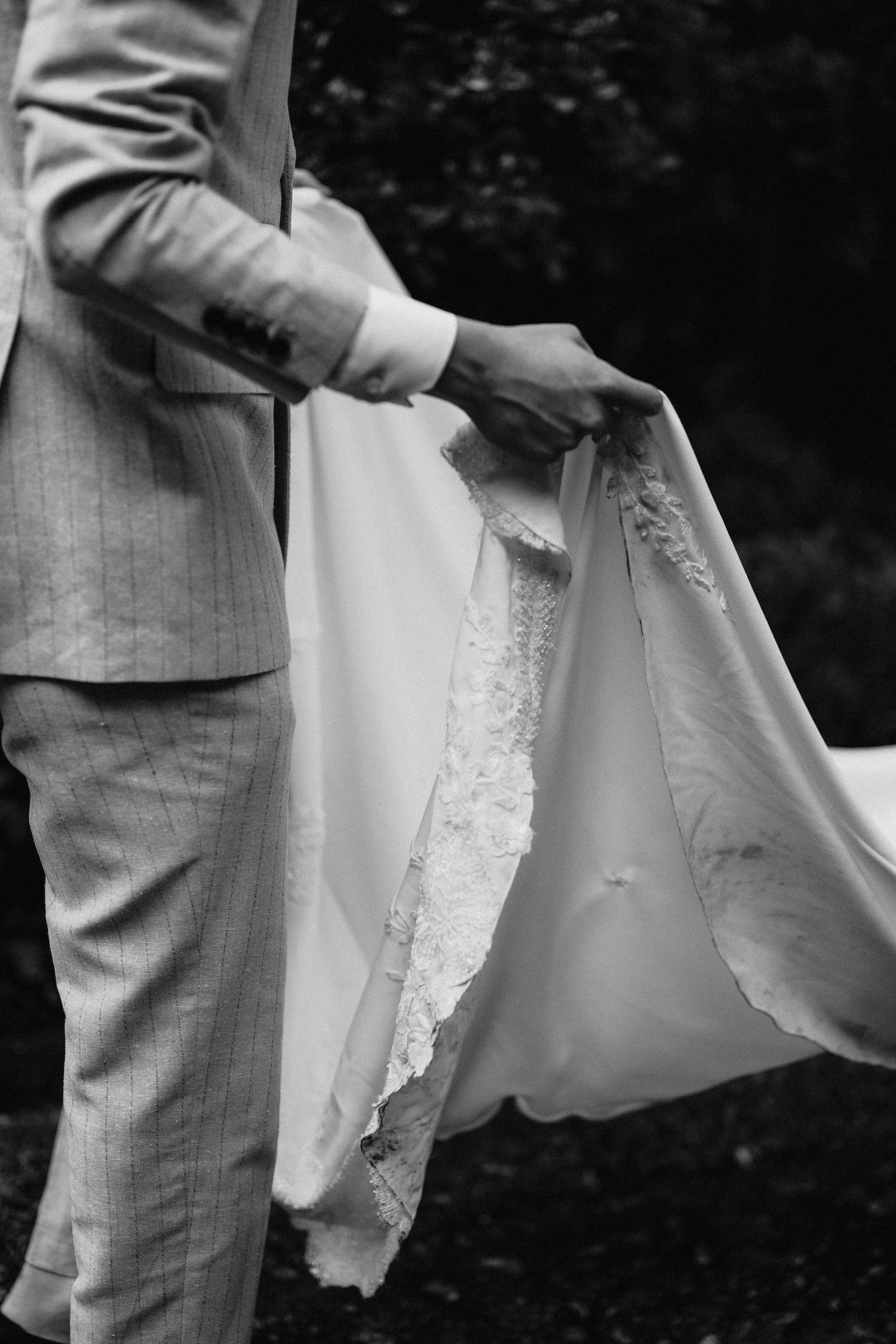Groom in pinstripe suit gently holds the train of brides lace wedding gown in black and white photo.