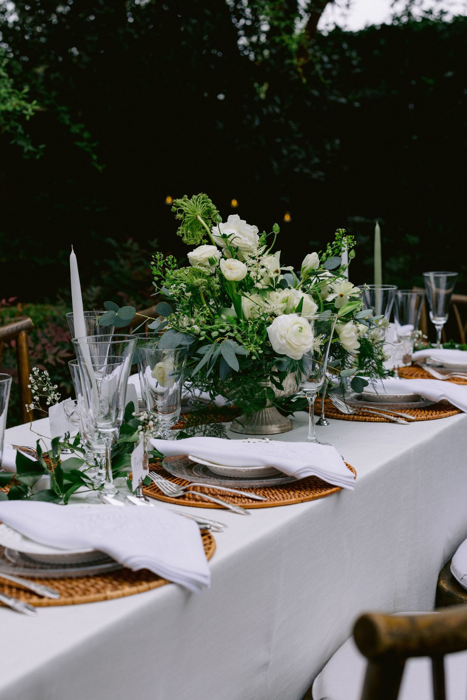 Outdoor wedding reception table with white roses centerpiece, green candles, wicker chargers, and white linen tablecloth.