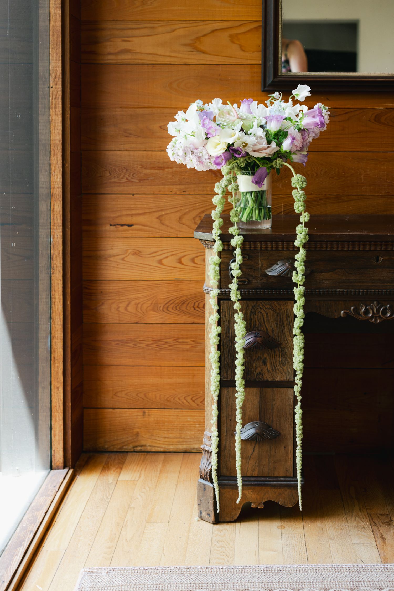 Elegant floral bouquet with cascading greenery displayed on a vintage wooden pedestal stand against warm wood-paneled wall.