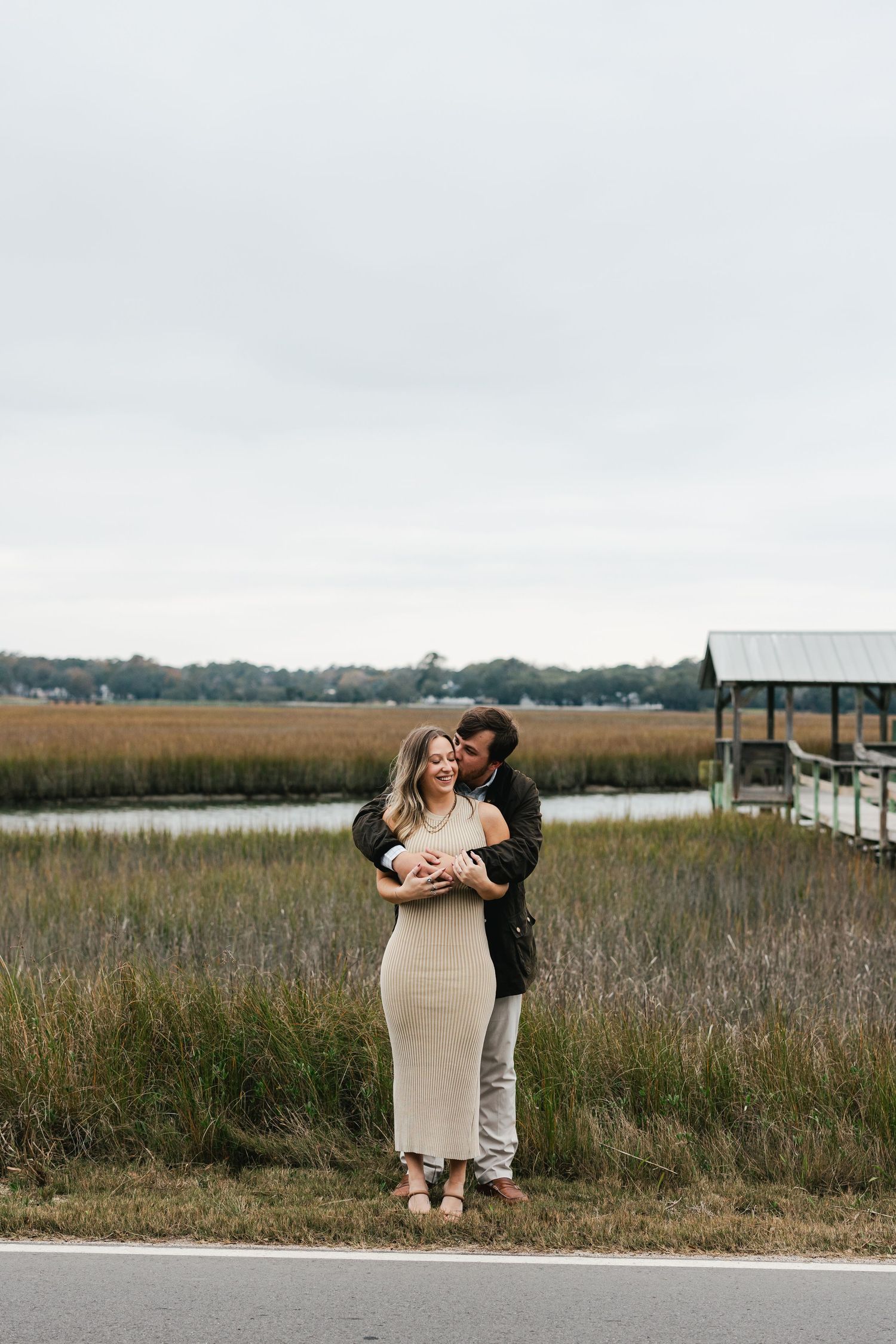 Couple embracing near a coastal marsh with tall grasses, a dock, and overcast skies in the background.