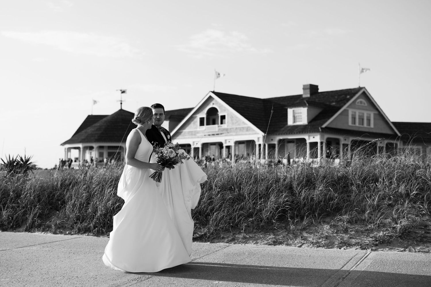 Black and white photo of a couple in wedding attire posing in front of a large coastal house on their wedding day.