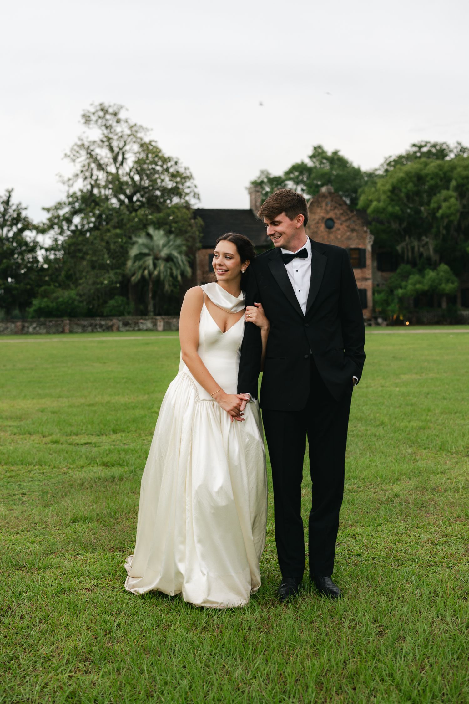 Bride in white gown and groom in black tuxedo stand together on green lawn with historic ruins backdrop.